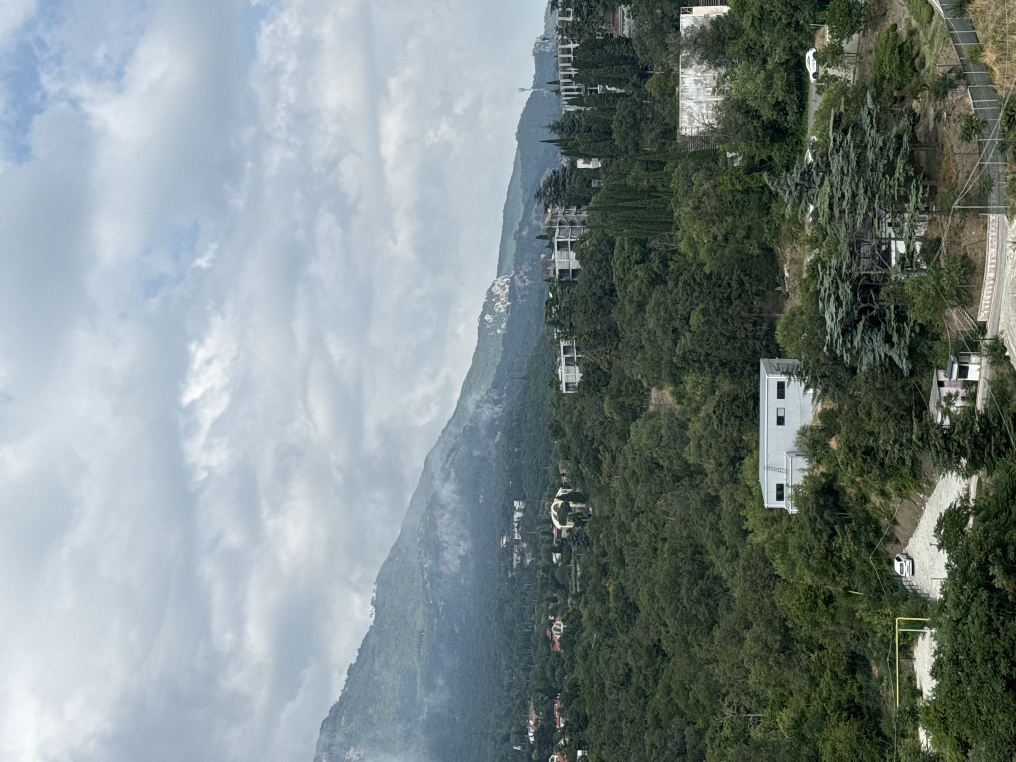 The image shows a mountainous landscape with a town nestled in the foothills, likely in the city of Yalta, Ukraine. The sky is overcast with clouds. Lush green trees cover the hillsides, with several buildings scattered throughout the scene. In the foreground, there is a modern, rectangular building with windows and a ladder on its exterior. A white car is parked in a gravel lot near this building, beside a yellow gas pipe. Further up the hill, more houses and apartment buildings are visible, some appearing to be under construction. Tall cypress trees are interspersed among the buildings. The overall impression is of a residential area built into a wooded, hilly terrain.