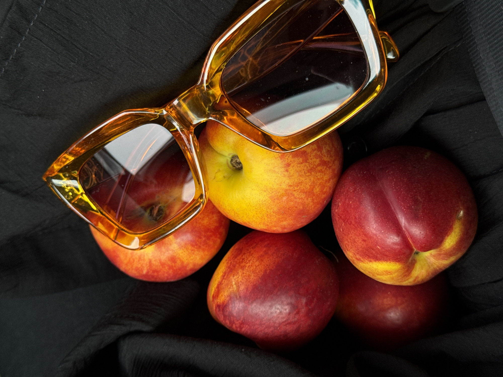 The image shows a still life arrangement of fruit and sunglasses on a dark background, possibly cloth. There are five nectarines or peaches, ranging in color from yellow to deep red, grouped together. On top of them rests a pair of amber-framed sunglasses. The setting appears to be indoors. The image provides no information to indicate that this is in Bersut-Sukache, Russia.