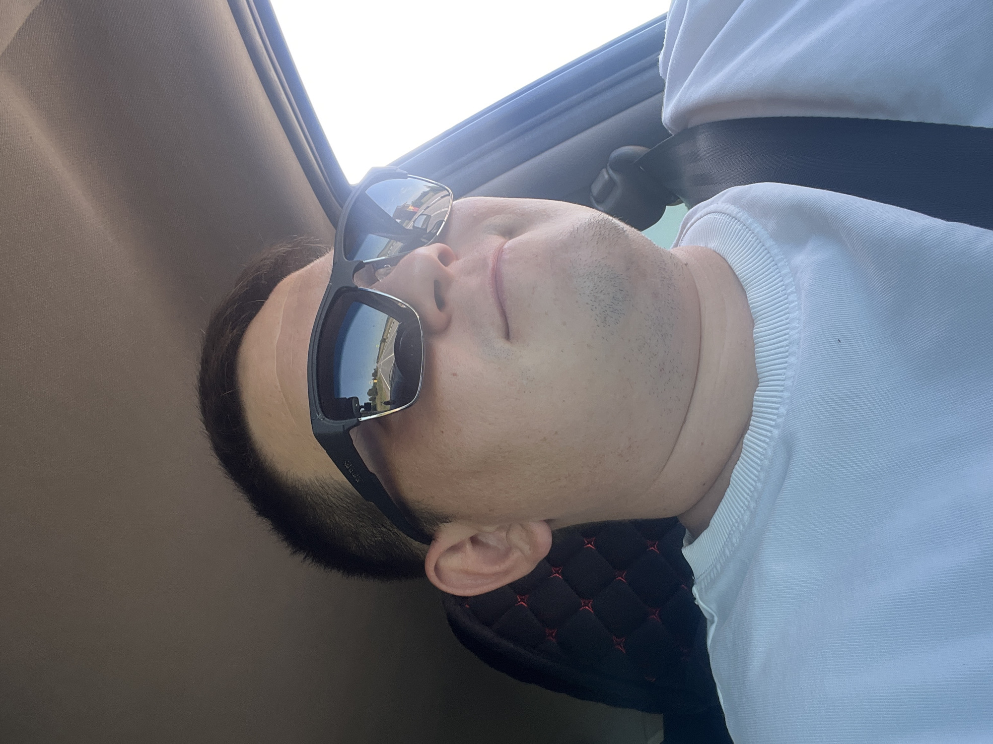 A medium shot captures a person wearing sunglasses and a white t-shirt while seated in a vehicle. The person is looking slightly off-camera to the right. The sunglasses reflect a road scene with a truck and a clear sky. The interior of the car is visible, including a black seat cover with red accents and a seatbelt. The overall lighting suggests it is daytime. The image is not from Maloe Churashevo, Russia.