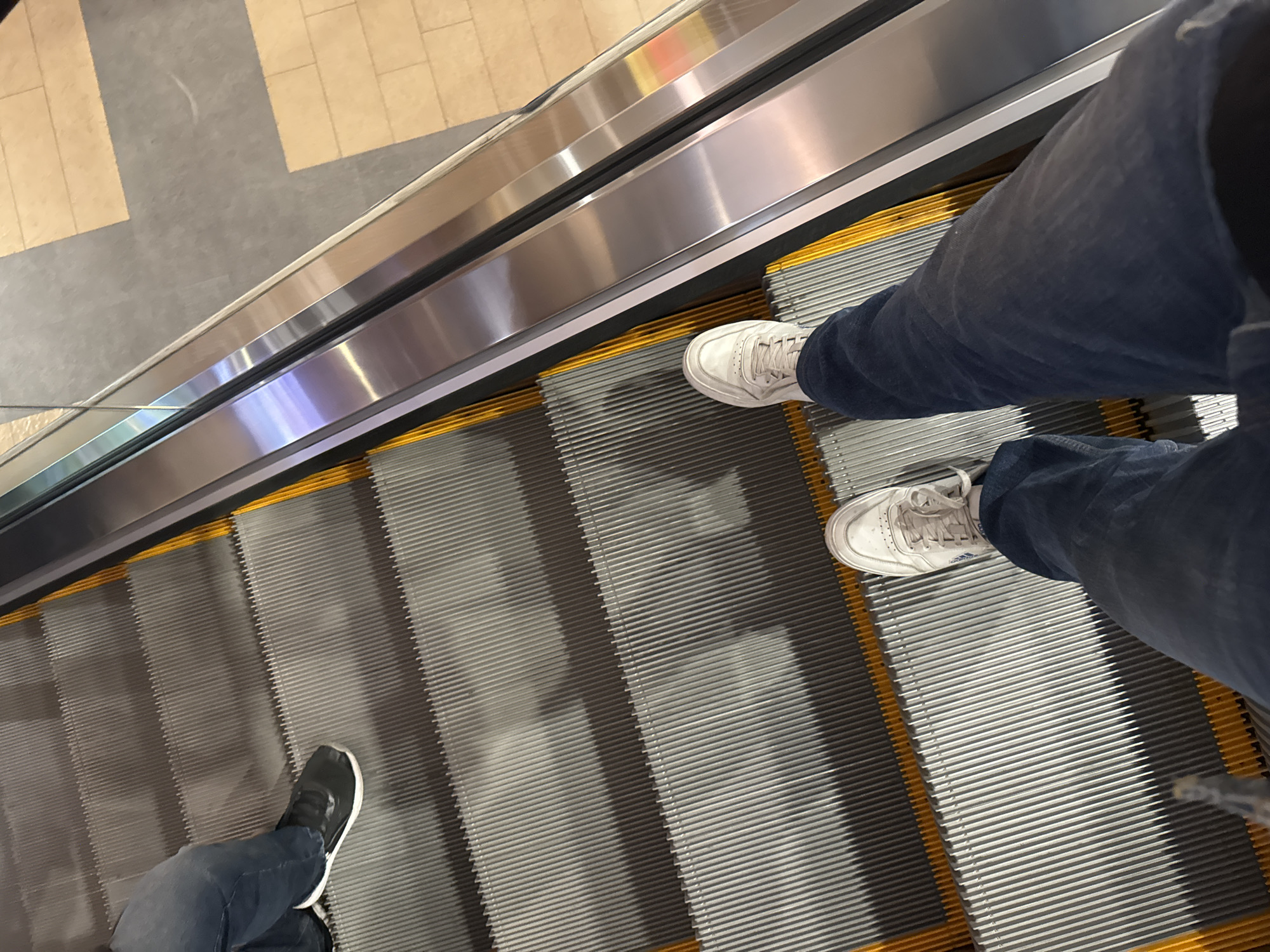 The image shows a view from above looking down on an escalator with two people riding it. The escalator has a metallic silver frame with yellow borders along the edges of the moving steps. One person is wearing blue jeans and white sneakers. The second person is wearing blue jeans and black sneakers with white trim. The background shows a beige and gray tiled floor. The location is likely a commercial building, potentially a shopping mall. There is no explicit information to identify the location in Puebla, Mexico.