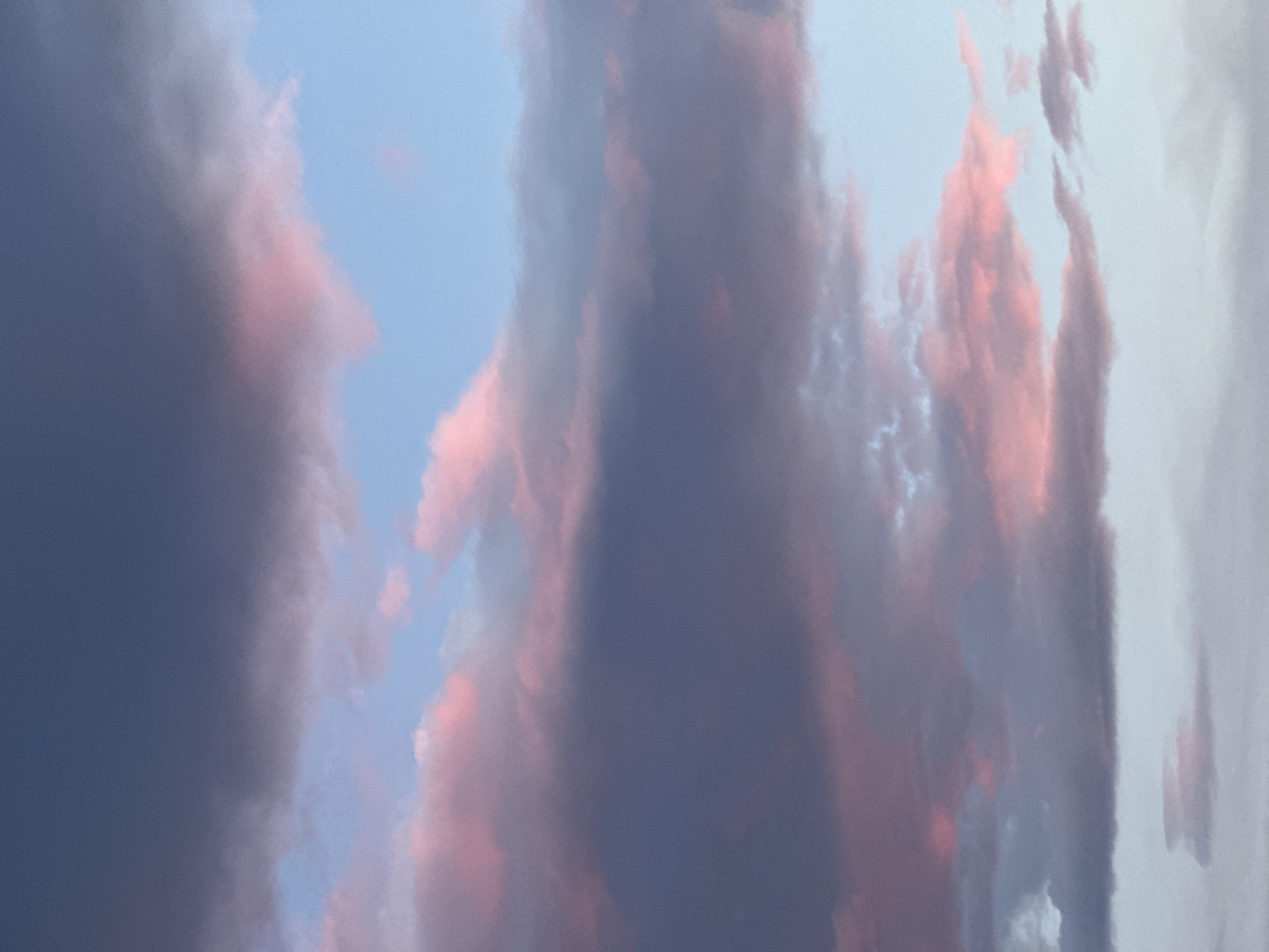 The image depicts a sky filled with clouds during a sunset or sunrise. The clouds are a mix of colors, including dark blue-gray, light blue, and pink. The pink color is likely due to the sun's rays reflecting off the clouds. The image does not depict any specific location or detail indicating that it was taken in Bar, Montenegro. The image is focused on the sky and the clouds. There are no people, subjects, objects, actions, or interactions visible.