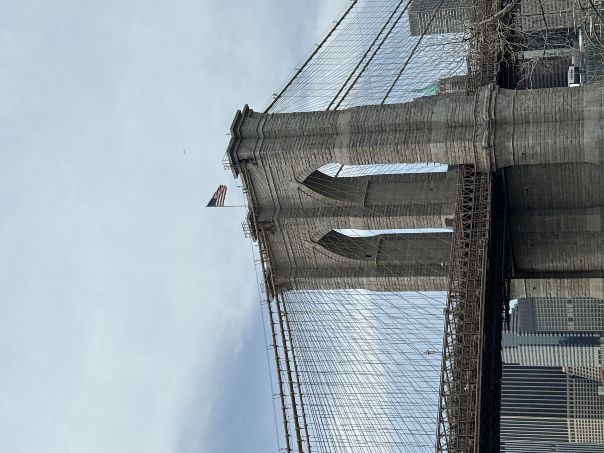 The image captures a section of the Brooklyn Bridge in New York City, USA. The gray stone tower stands prominently, featuring arched openings. An American flag flutters from the tower's top. A complex network of steel cables spans out from the tower. The background includes views of city buildings and a pale sky. The photo is taken from a low angle, looking upwards.