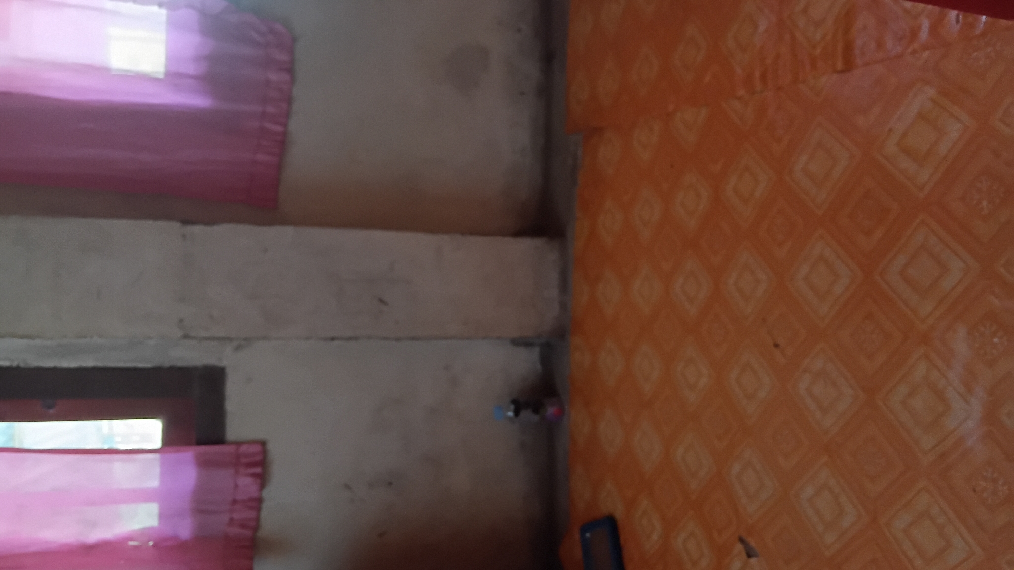 The image depicts an interior scene, likely a room, with a focus on the lower half of the space. The floor is covered with orange tiles featuring a repeating diamond pattern with lighter colored square inlays. In the foreground left, a blue rectangular object, possibly a tablet or a book, rests on the floor. Further back, near the wall, a small, multi-colored object, potentially a toy or a decorative item, is visible. The walls are a neutral, textured color, and a concrete pillar stands prominently in the center of the image, dividing the space. The upper portion of the image shows portions of two windows, each draped with translucent pink curtains that allow light to filter through. The overall impression is of a simple, lived-in interior space. The image is not specific to Prabumulih, Indonesia, and lacks any contextual clues to place it in that location.