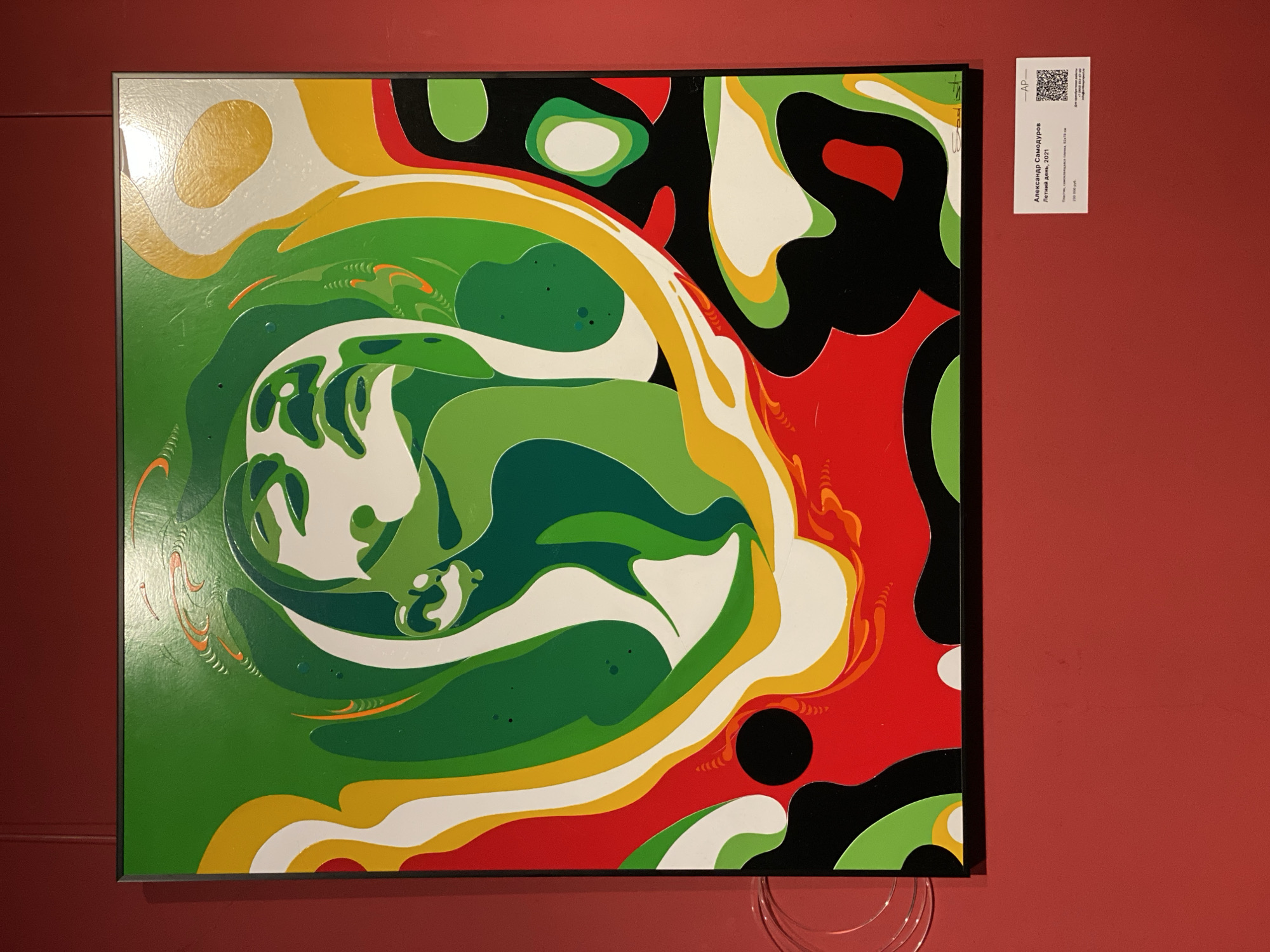 The image shows a brightly colored abstract painting hanging on a red wall. The painting is primarily composed of green, white, yellow, red, and black shapes, with a swirling pattern in the center. A small white tag is attached to the wall to the right of the painting, with some text visible, including the name "Александр Самодуров" (Aleksandr Samodurov). The label indicates that the painting is titled "Летний день, 2021" (Summer day, 2021) and the price is 250,000 rubles. Based on the artist's name and the use of the Cyrillic alphabet, the location is likely Moscow, Russia. A few transparent cords hang down below the painting.