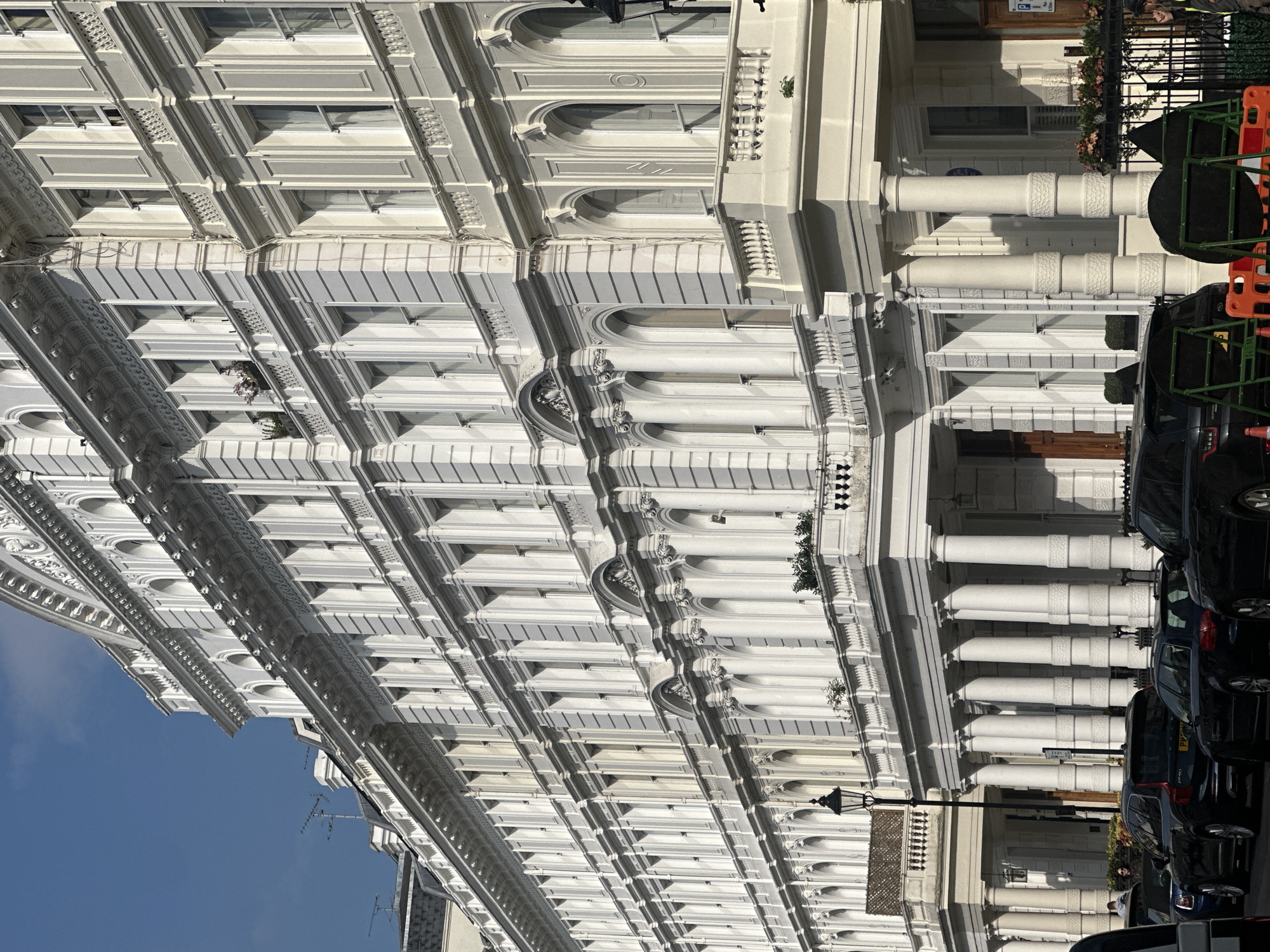 The image showcases a row of elegant, white-painted buildings in London, United Kingdom. The architectural style features ornate detailing, including columns, arched windows, and decorative molding. The buildings rise several stories, creating a uniform and imposing streetscape. A row of parked cars lines the street, partially obscuring the ground level of the buildings. A lamppost stands along the sidewalk. The sky is visible in the upper left corner, exhibiting a clear blue hue.