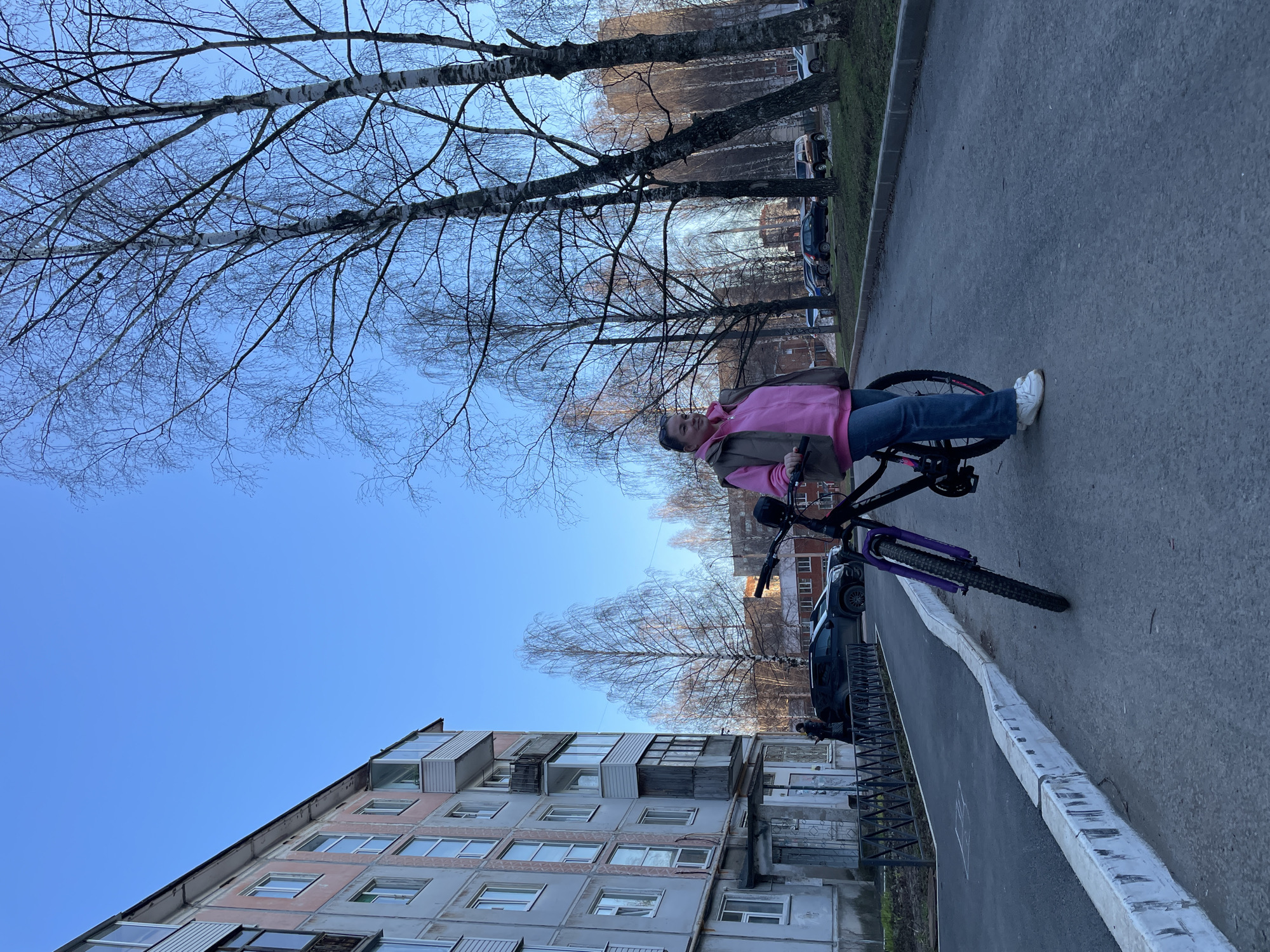 A street scene in Izhevsk, Russia, shows a woman on a purple bicycle on a sidewalk next to a multi-story apartment building. The woman wears a pink hooded sweatshirt, a brown jacket, and blue jeans, with white shoes. The bicycle has black handlebars and a black frame. A gray curb separates the sidewalk from a gray asphalt road with parked cars and trees in the background. The apartment building is gray with multiple windows. Bare trees are visible against a blue sky.
