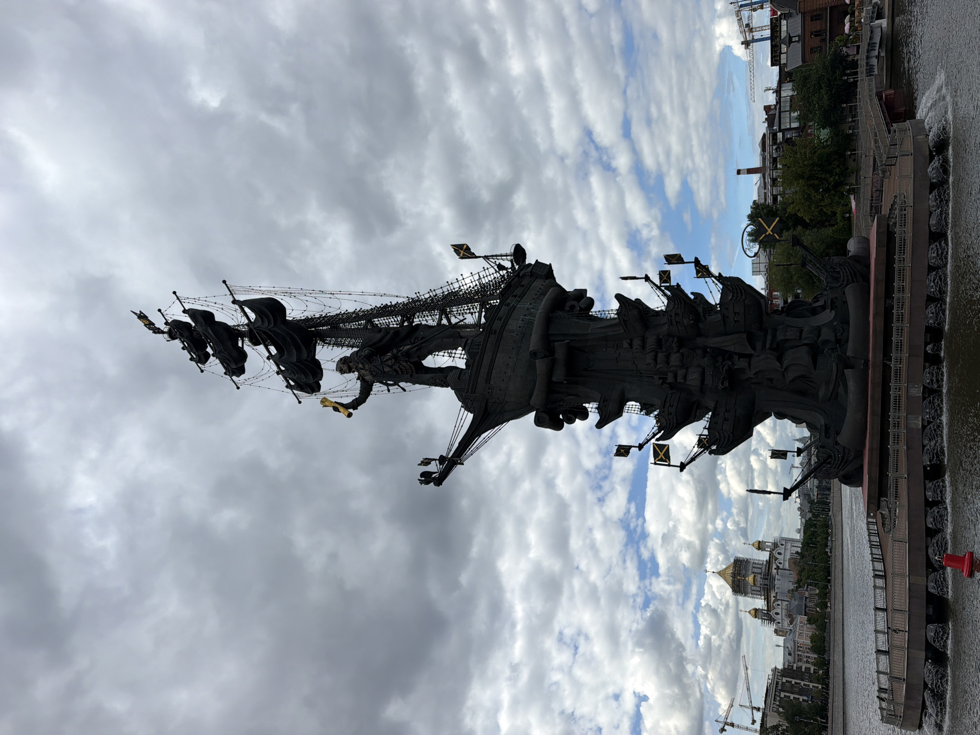 A vertical medium-shot photograph depicts the Peter the Great Statue in Moscow, Russia, captured under a cloudy sky. The statue, dominating the foreground, is constructed in dark metal and depicts a tall ship with various figures and flags. The ship's rigging is also visible.
In the background, the skyline of Moscow is partially visible. The domes of a church are apparent, as well as a series of modern buildings and construction cranes. The water of the Moscow River is seen along the edge of the photo, with the base of the statue partially submerged.
