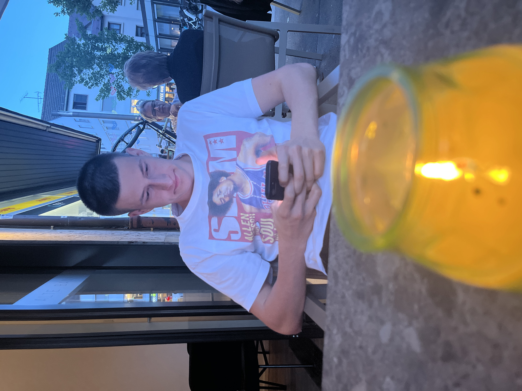A young man is seated at an outdoor table, looking at his smartphone. He's wearing a white t-shirt with a graphic featuring Allen Iverson. To the left, a storefront with glass windows is visible, and in the background, people are sitting and cycling. A yellow light source is in the right foreground. The location is a city of Unknown, Unknown.