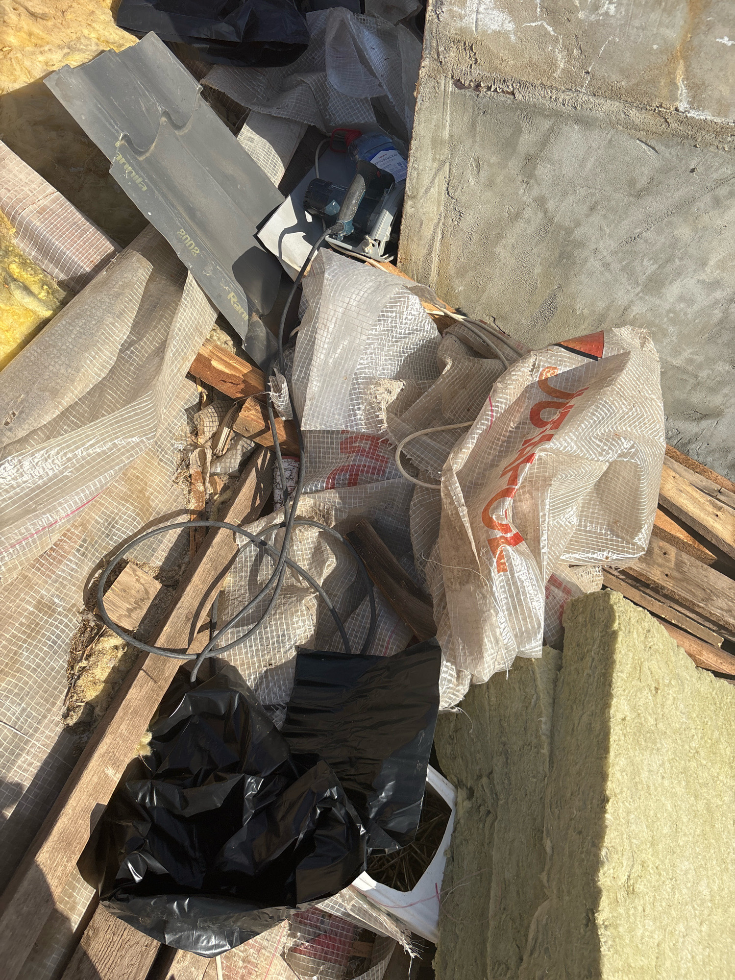 The image displays a pile of construction debris, including what appears to be insulation material, plastic sheeting, wooden planks, and a black trash bag. A gray roof tile with text partially visible, possibly including "2002" and "Marquinha", is also present. In the upper right quadrant, a concrete wall is visible. The scene is lit by sunlight. No people are visible in the image. The debris is located outdoors, likely in a construction or renovation area. The location is identified as Zhavoronki, Russia.