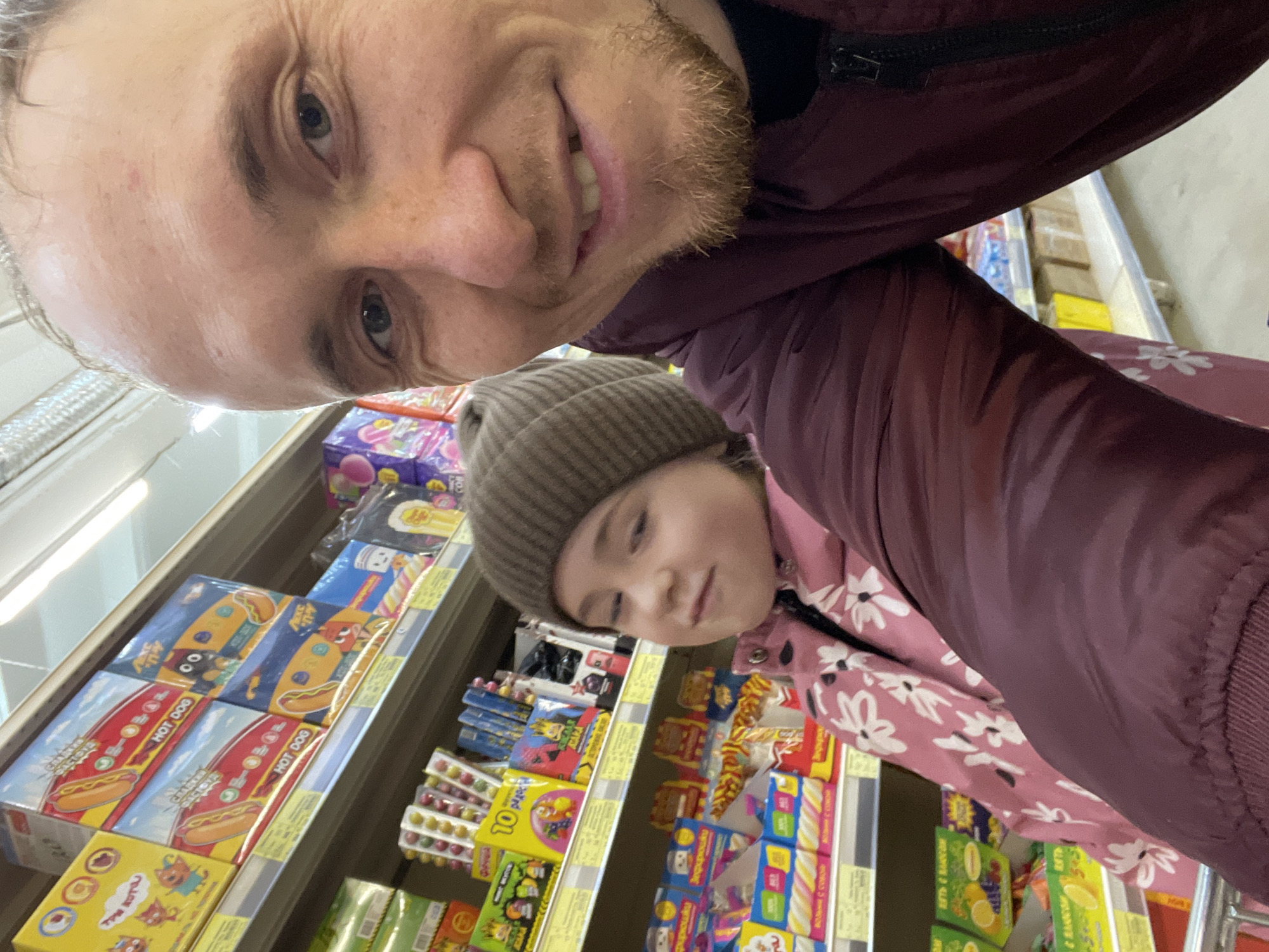 A selfie taken inside a store shows an adult male smiling in the upper portion of the frame, with a young girl wearing a beanie positioned below him. The man is wearing a maroon jacket. The girl is wearing a pink jacket with a floral pattern and a brown beanie. They are standing in front of a shelf stocked with various products, including candy and snacks. Notably, boxes labeled "HOT DOG" are visible on the shelf. The products are written in Russian, suggesting the location is in a Russian-speaking area. The store could be in Sarapul, Russia.