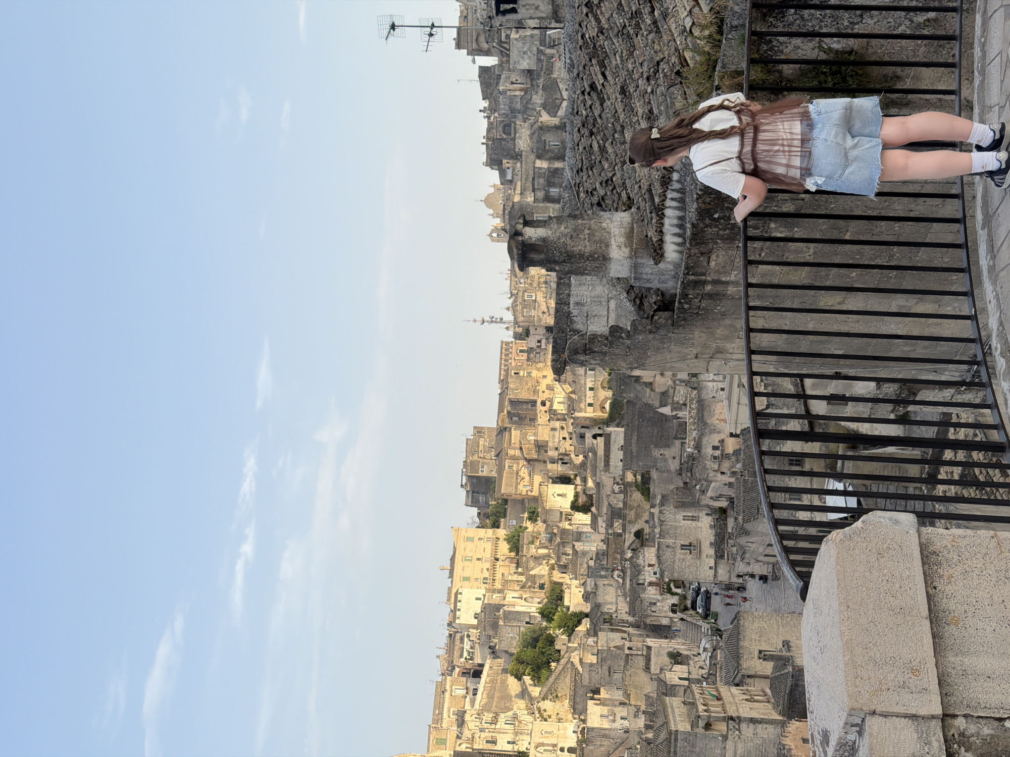 A young woman with long, brown hair, wearing a white t-shirt, a sheer brown overlay top, and denim shorts, stands with her back to the camera, looking out at the city of Matera, Italy. She is positioned on a balcony with a black metal railing, with a stone wall to her left. The background is a panoramic view of the historic Sassi di Matera, characterized by ancient cave dwellings and stone buildings cascading down a hillside. The sky is a clear, light blue with a few wispy clouds. The overall lighting suggests it is late afternoon or early evening, with warm tones illuminating the cityscape. Several people can be seen walking in the distance on a street below.
