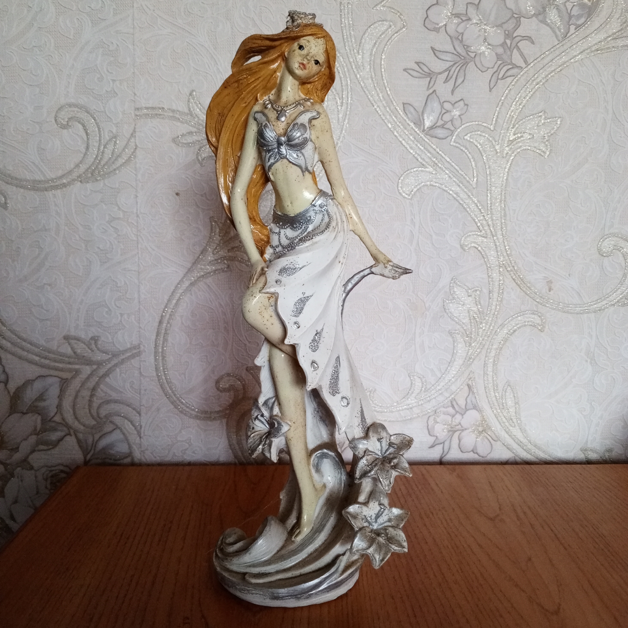 A decorative figurine of a woman stands on a wooden surface in front of a patterned wallpaper. The figurine is slender with long, flowing auburn hair and is adorned in a white, flowing dress with silver accents, including a butterfly-shaped top and intricate jewelry. The base of the figurine is sculpted to resemble waves and flowers. The wallpaper behind the figurine features a light-colored, floral damask pattern with subtle shimmering elements. The wooden surface appears to be a tabletop or shelf. The scene is indoors, likely within a domestic setting. The specific location in Sayanogorsk, Russia, cannot be determined from the visual information provided.