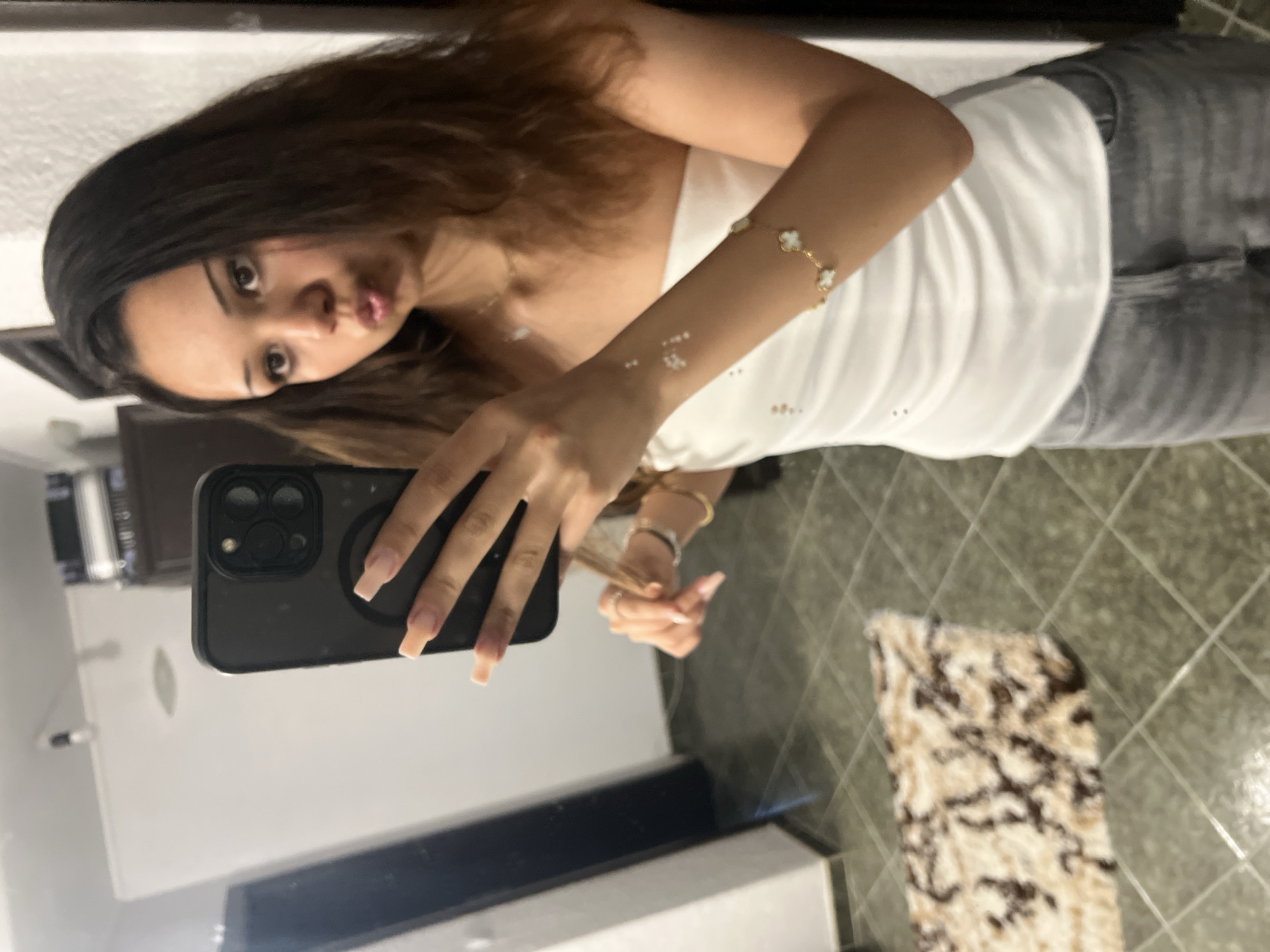 A young woman with long dark brown hair and a light complexion captures a mirror selfie indoors. She wears a white strapless top and grey jeans. Her left hand holds a dark-colored smartphone, featuring a prominent camera array and a circular magnetic attachment on the back, oriented vertically for the photo. Her fingernails are long and painted a light, neutral color. Her right hand is near her hair, and she wears a gold bracelet with white clover-shaped charms on her right wrist, along with small white dot-like designs or embellishments on her right forearm. A thin gold necklace is visible around her neck. She pouts her lips towards the camera. The reflection reveals a room with white walls, a dark door or frame on the left, and a dark green/grey tiled floor with a diamond pattern. A beige and brown patterned rug is partially visible on the floor to the left. Overhead, a light fixture is reflected. In the upper right background, a dark piece of furniture or cabinet is visible. The location is an unspecified room within a private residence in the city of Unknown, Unknown.