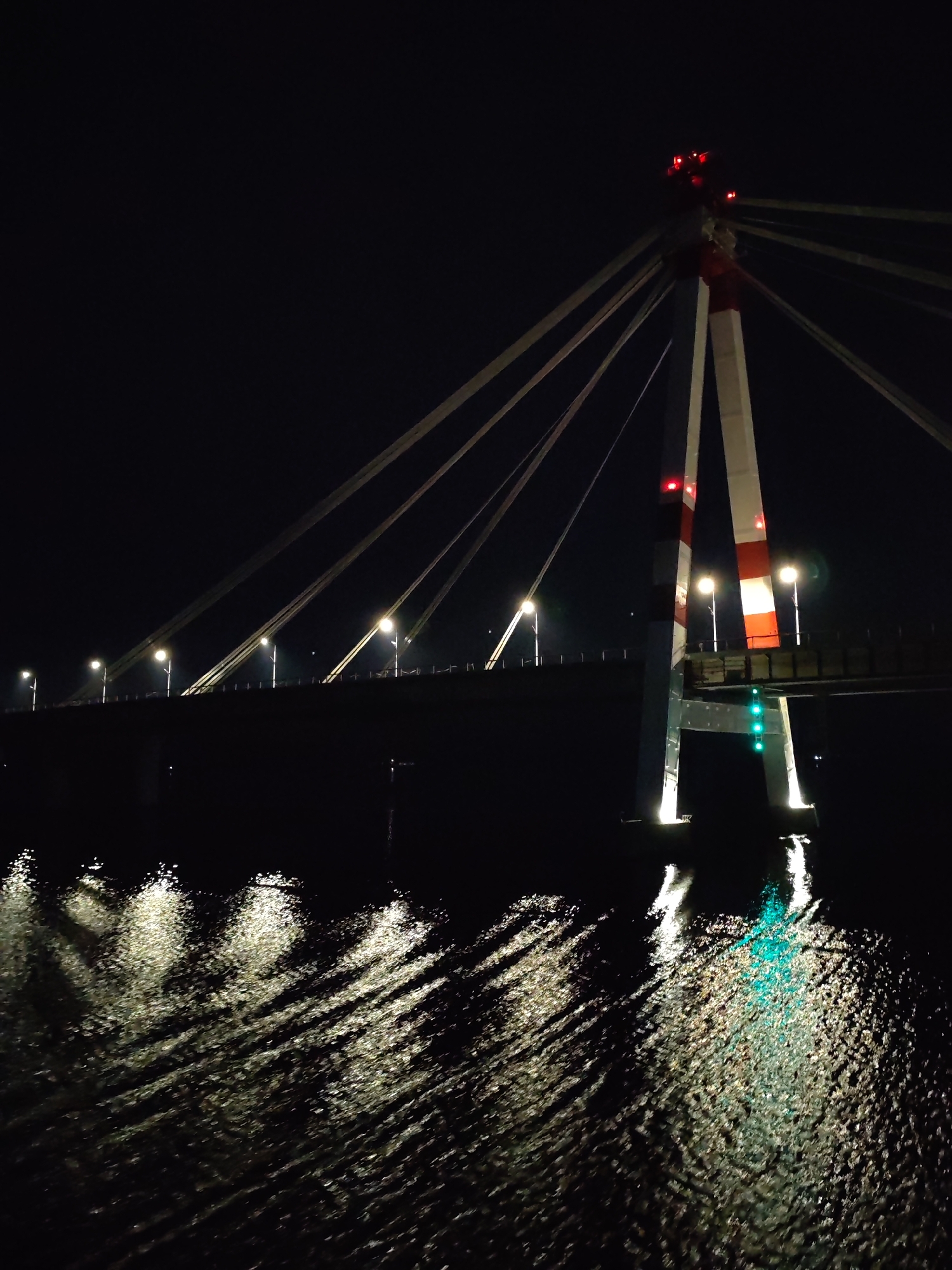 A nighttime photograph depicts a bridge over water. The bridge is a cable-stayed structure, featuring a central tower with red and white stripes. Cables extend from the tower to the bridge deck. Several streetlights illuminate the bridge deck. The water below reflects the bridge's lights, creating patterns on the surface. Three green lights are visible below the tower on the right side of the image. The sky is dark, with the bridge lights being the primary source of illumination. Location is Unknown, Unknown.