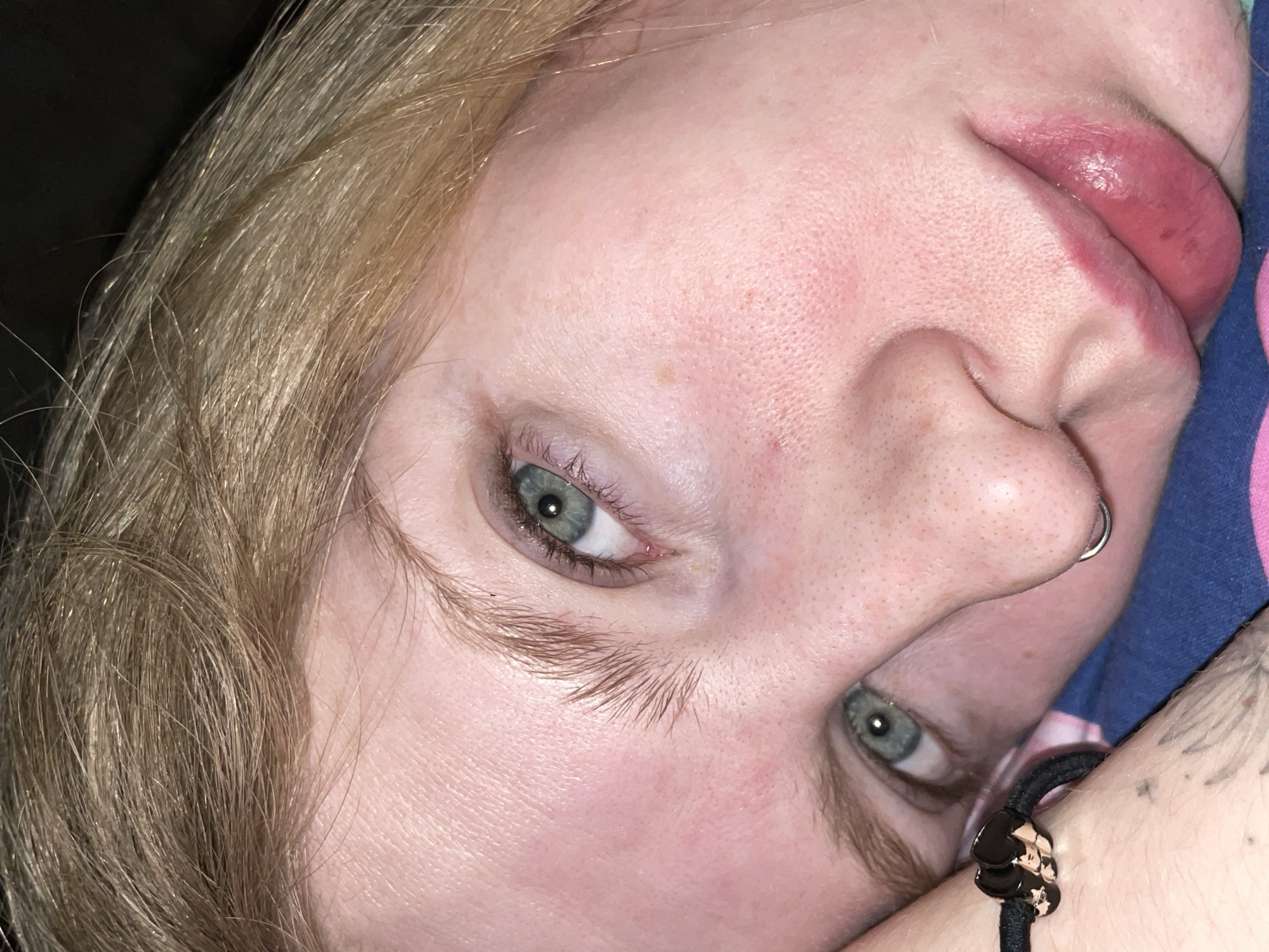 The image is a close-up of a person's face. The person has light blonde hair, fair skin, and light green/blue eyes. They have a nose piercing on the left nostril. The person is wearing a dark bracelet with a small black skull-shaped charm. The lips appear swollen. The background is out of focus, but some fabric with blue and pink colors is visible. There is no indication of Izhevsk, Russia in this image.