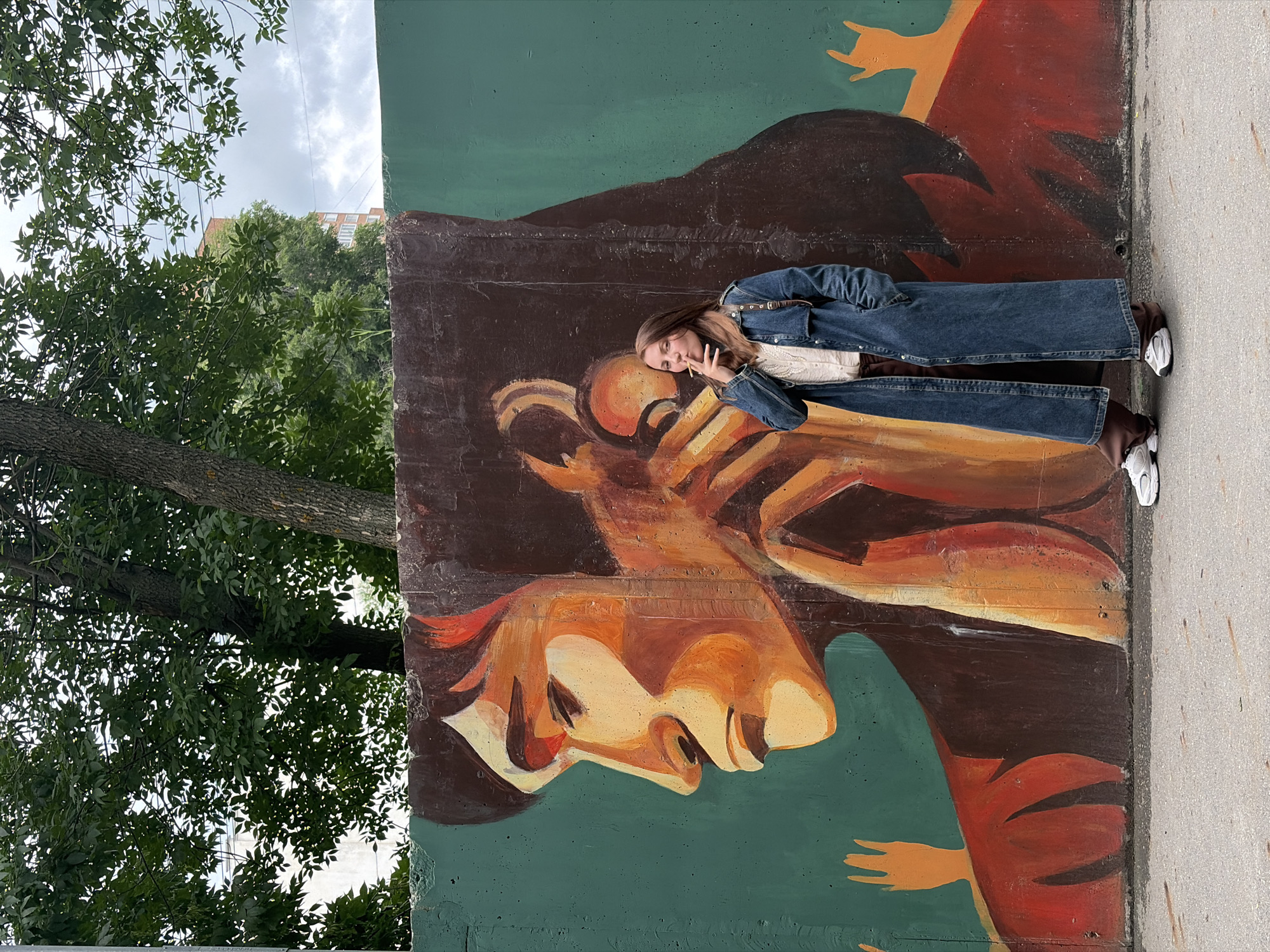 A young woman stands in front of a mural in Izhevsk, Russia. She is wearing a long denim jacket over a white blouse and brown pants, with white athletic shoes. She is holding a cigarette to her lips and appears to be smoking. The mural depicts a profile of a person with dark hair, rendered in warm tones of orange, yellow, and brown against a teal background. Two orange hands are visible on either side of the main figure in the mural. The woman is standing on a paved surface, with trees and a building visible in the background. The sky is overcast.