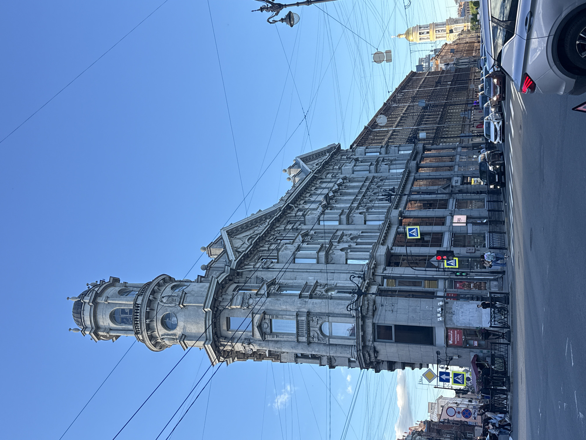 This is a daylight, eye-level, full shot of a prominent, angular building located at a street intersection in Saint Petersburg, Russia. The building, a mixture of architectural styles, features a distinctive rounded tower with a domed roof and decorative elements at its apex. The facade is ornate, with numerous windows, balconies, and intricate stonework. Directly in front of the building, a street corner is visible with pedestrian crossings, traffic lights, and street signs. Several people are walking on the sidewalk and crossing the street. The street is active with vehicular traffic, including cars and a bus. Overhead, a complex network of telephone and electrical wires crisscrosses the sky. Further down the street, other buildings can be seen, including a distant church with a golden dome. The sky is clear and blue.