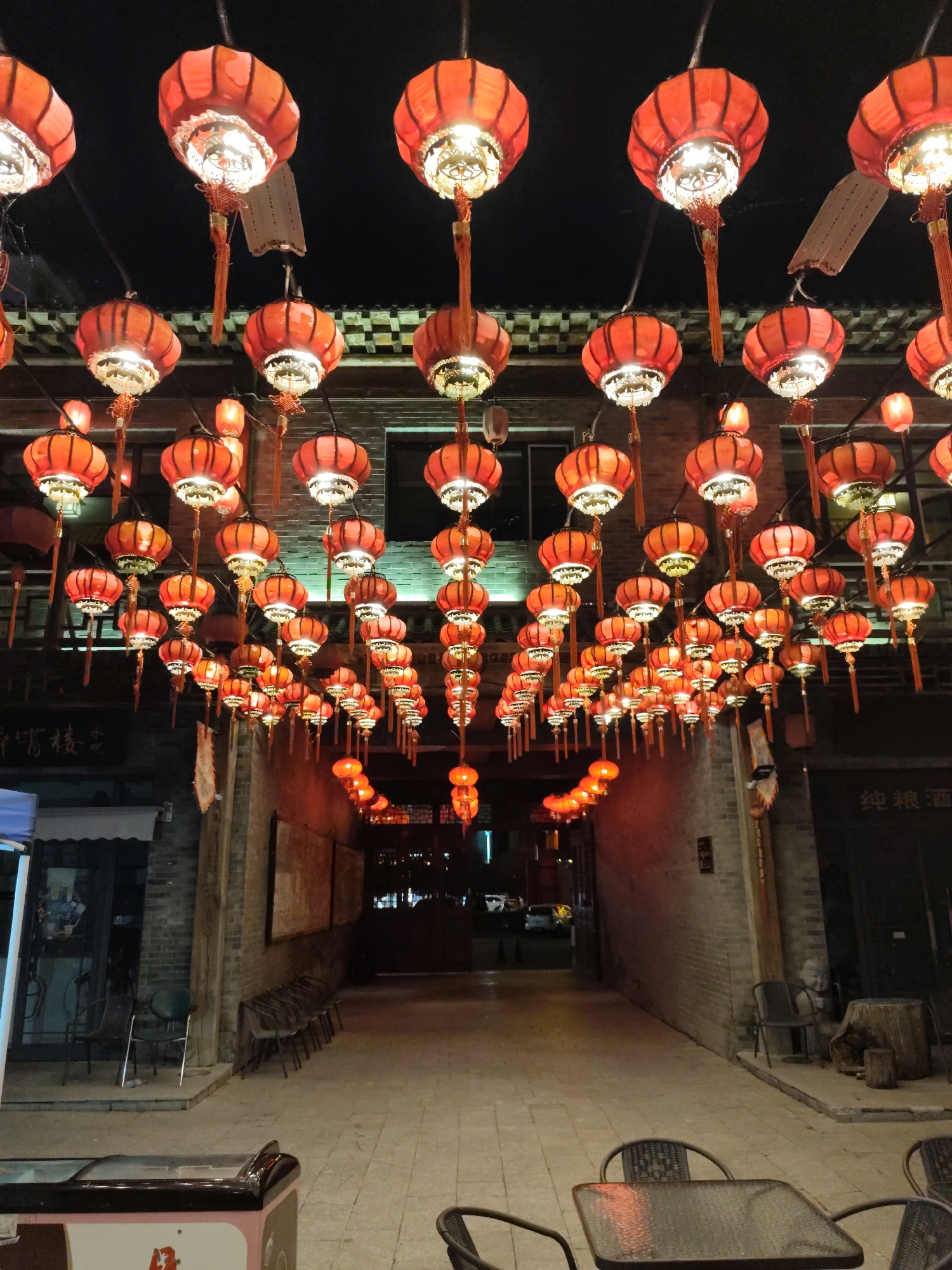A street scene in Lindian, China, is illuminated by dozens of red, glowing lanterns hanging overhead. The lanterns are suspended from wires strung across a narrow alleyway lined with brick buildings. The alley opens into a wider area with tables and chairs set up for outdoor dining, suggesting a restaurant or food establishment. The ground is paved with light-colored cobblestones. In the foreground, a table with chairs is visible, and to the left, a glass-fronted refrigerator or display case stands. The overall atmosphere is festive and warm, likely during an evening or night setting.