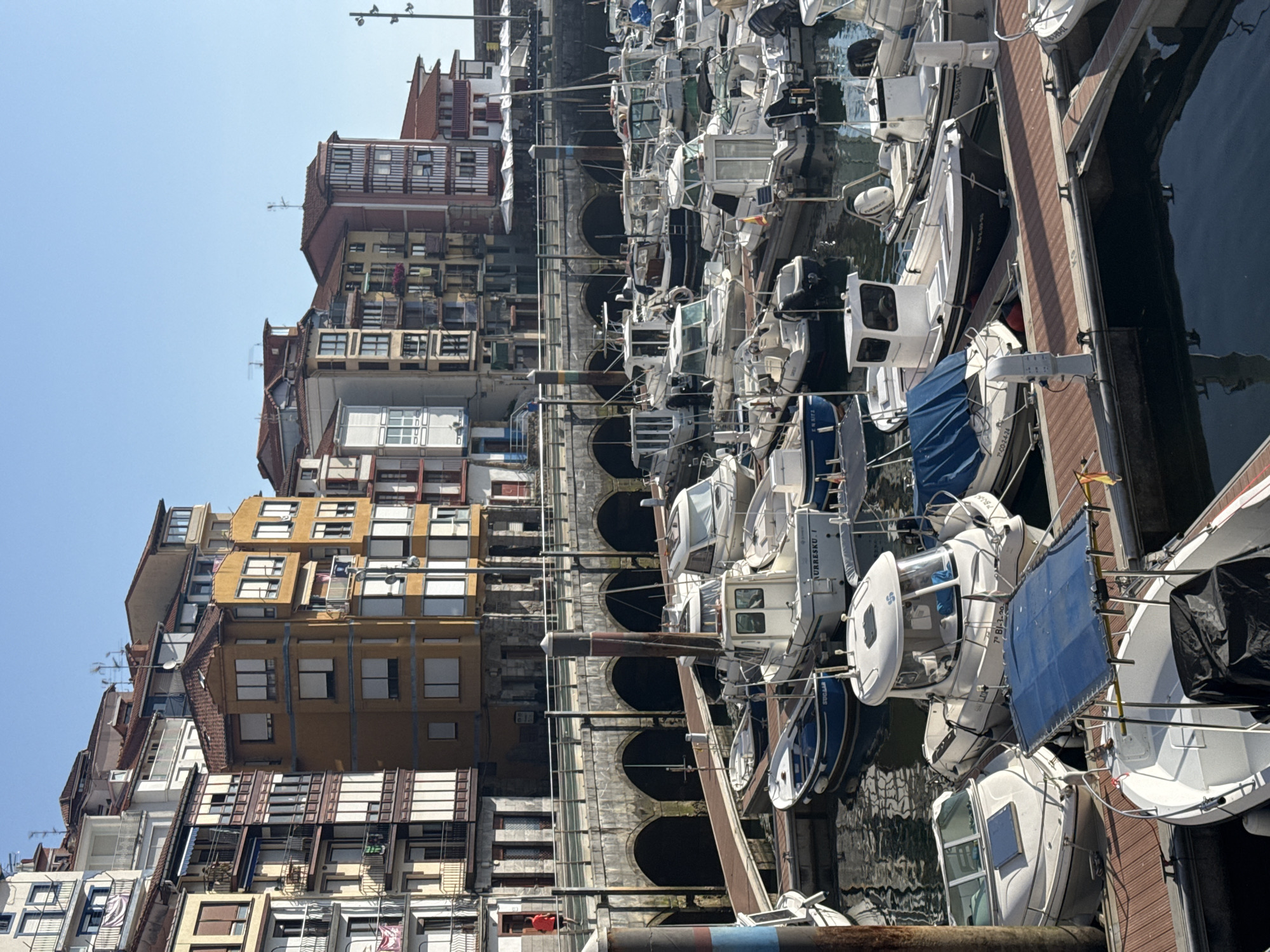 A wide shot captures a sunlit harbor in Bermeo, Spain, with colorful buildings lining the waterfront. The scene is dominated by a marina filled with numerous boats of various sizes, docked and bobbing on the calm water. The buildings are multi-story structures with distinct architectural styles, featuring balconies, windows, and tiled roofs in shades of yellow, orange, and white. The sky is a clear, bright blue, indicating fair weather. In the foreground, wooden docks provide walkways for accessing the boats. One boat in the center has the name "AURRESKU I" visible on its side. Another blue boat has "SUGAR" written on it. The overall impression is of a peaceful, picturesque coastal town.