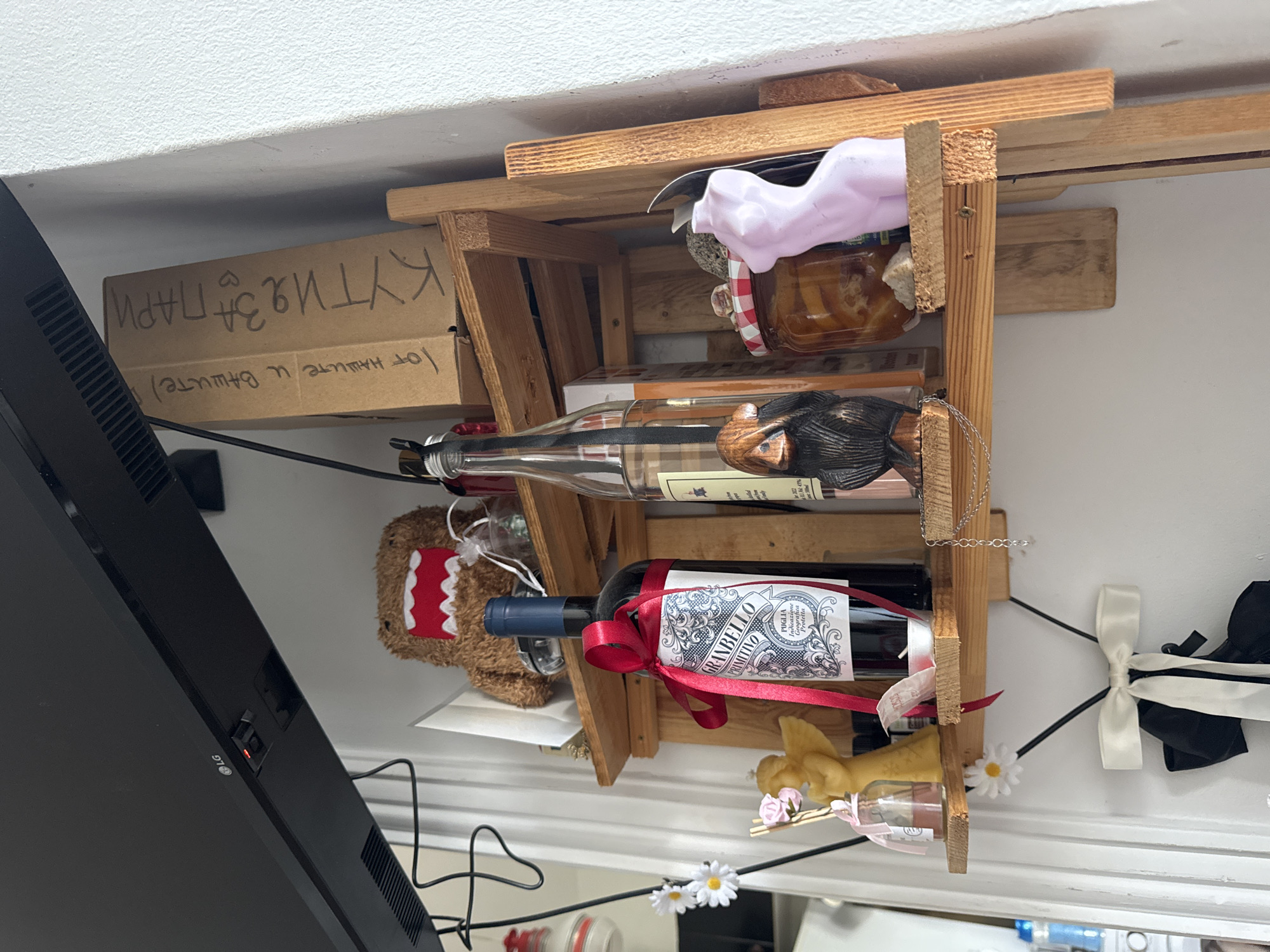 The image shows a close-up of a wooden shelf holding various items, with a black LG monitor in the foreground. The shelf is made of unfinished wood and appears to be home-built, with rough edges and visible nails.
On the top shelf, there's a glass jar with a red and white checkered lid, containing a dark-colored substance, possibly jam or honey. Next to it, a pink, translucent object, possibly a plastic mold or figurine, is also present.
The middle shelf holds two bottles. The top bottle is clear glass with a dark liquid and a label that is partially visible. The bottom bottle is a dark glass wine bottle with a prominent red ribbon tied around its neck and a detailed label that reads "GRANBELLO PRIMITIVO PUGLIA Indicazione Geografica Protetta".
On the bottom shelf, there is a small, brown teddy bear with a red mouth, and a small glass bottle with a yellow stopper and a pink bow. Next to it, a yellow wax figurine, possibly an angel, is visible with pink flowers.
To the left of the shelf, a cardboard box is visible with Cyrillic writing that translates to "BOX FOR MONEY" and "From our and yours." The LG monitor in the foreground has a power button with a red indicator light.
The overall scene suggests a domestic setting, likely a home or a small shop, with a mix of decorative and functional items. The lighting appears to be artificial, coming from above and to the right. The image is taken with a slight upward tilt, implying the shelf is mounted higher up.