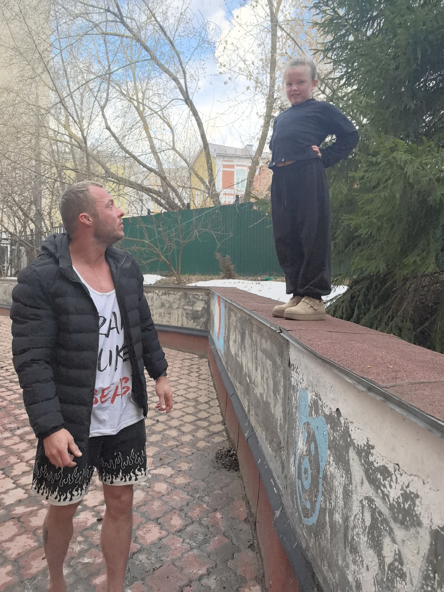 A medium shot captures a man and a young girl in an outdoor urban setting. The man, dressed in a black puffer jacket over a white tank top with red lettering, and black shorts with a white flame pattern, stands on a wet, brick-paved ground. He gazes upward toward the girl, who is standing on a low, brick-topped wall with painted graffiti on its side. She wears a black long-sleeved top, black pants, and beige shoes.
The background includes deciduous trees with sparse foliage, a green wooden fence, a building with white window frames, and a conifer tree to the right of the girl. The sky is visible above the trees, with patches of blue and white clouds. The location is likely in Nizhny Novgorod, Russia.