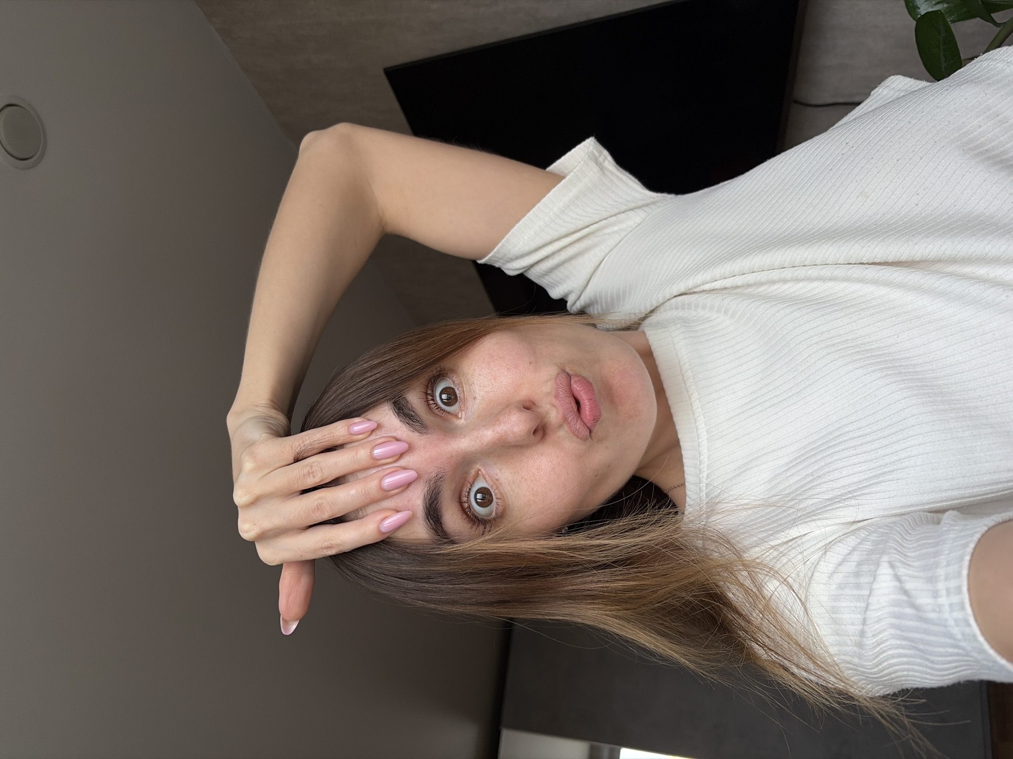 The image is a vertical selfie of a young woman, rotated 90 degrees clockwise, depicting her in a playful or surprised pose. She is looking directly at the camera with widely opened brown eyes and puckered lips. Her light brown hair, with subtle highlights, frames her face, and visible freckles dot her cheeks and nose. Her right hand is raised, fingers spread, touching her forehead, revealing long fingernails painted a light pink shade.
She is wearing a light-colored, possibly off-white or cream, ribbed short-sleeved top. The setting appears to be indoors, likely a home, with a dark flat-screen television or monitor visible in the background behind her upper body. A light grey wall with a circular ceiling vent or light fixture is visible on the left side of the frame, and a touch of greenery from a plant can be seen in the upper right corner. The lighting suggests daytime. There is no visible text. The image's location context is Izhevsk, Russia, but this is not visually verifiable within the frame.