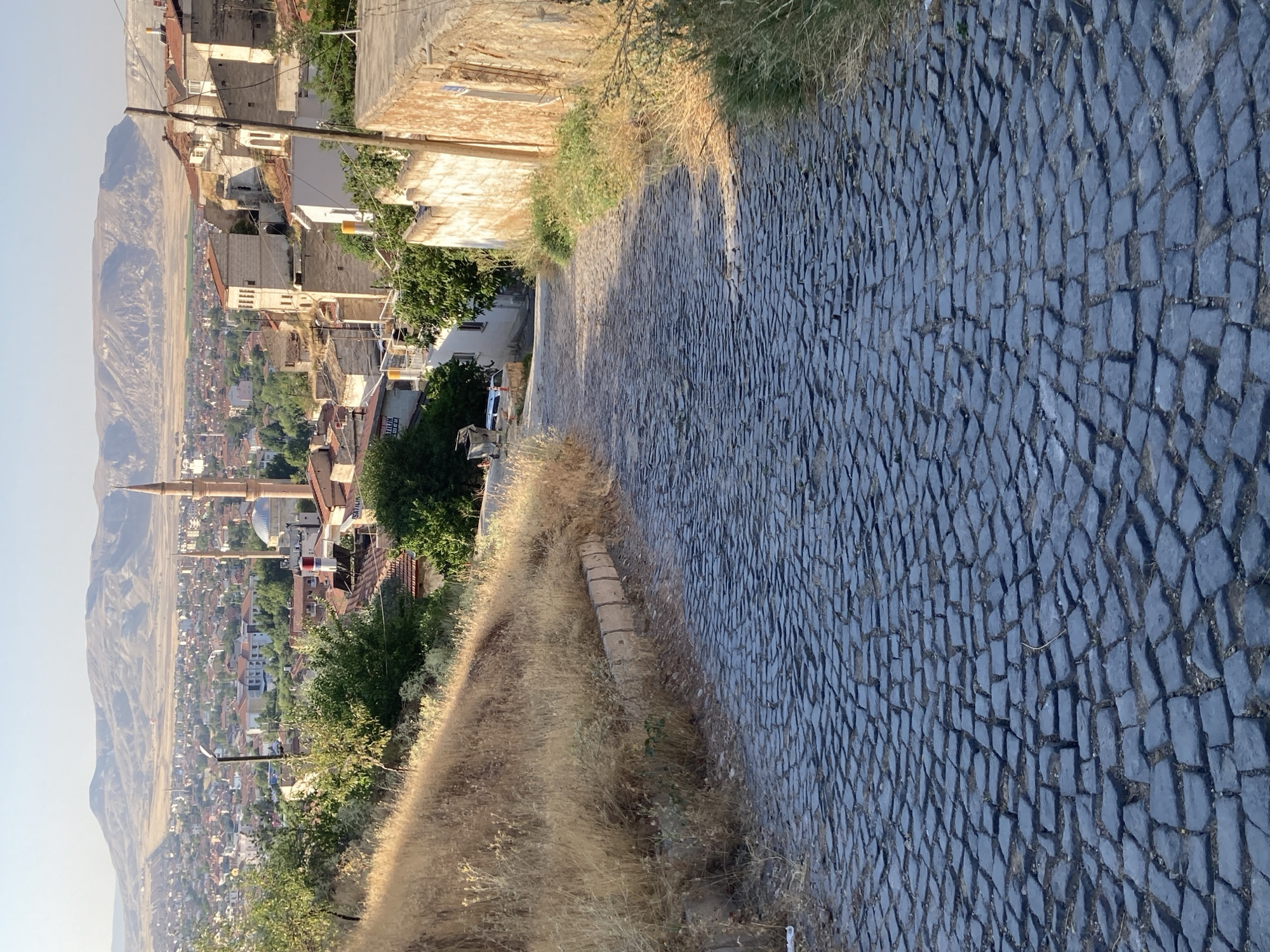 This image captures a downward sloping, cobblestone path in the city of Avanos İlçe Merkezi, Turkey. The path is paved with dark, irregular-shaped stones. On the left side of the path, a grassy embankment covered in dry, golden grass rises sharply. To the right, a weathered stone wall lines the path, with patches of green and dry vegetation growing around it. Further in the background, a sprawling city landscape unfolds, characterized by a dense collection of buildings with red-tiled roofs. A prominent mosque with a tall, slender minaret stands out amidst the urban sprawl. In the distance, rolling, arid hills form the backdrop under a clear, bright sky. The lighting suggests it is late afternoon or early morning, casting soft shadows and highlighting the texture of the cobblestones and the dry grass. There are no people visible in the image.