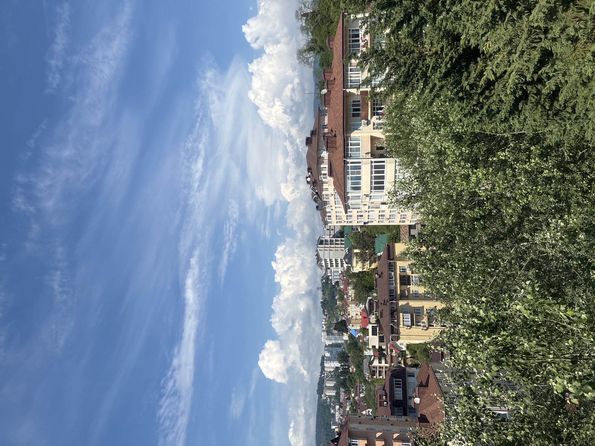 This image captures a view of the city of Sochi, Russia, characterized by a blend of residential buildings nestled amongst lush green foliage and against a backdrop of rolling hills under a bright blue sky dotted with white clouds.
The foreground is dominated by dense trees, their leaves appearing vibrant and full, suggesting a warm season. As the perspective shifts upwards, various buildings come into view. These range from multi-story apartment complexes with modern architecture and numerous windows, to smaller residential buildings with pitched, red-tiled roofs. The architectural styles suggest a developed urban area, possibly a residential neighborhood or a section of the city with mixed-use properties.
In the mid-ground and background, more buildings are visible, some appearing taller and more contemporary with lighter-colored facades, while others are smaller and closer to the ground. The hills in the far background are covered in greenery, indicative of a temperate climate and possibly proximity to mountainous terrain.
The sky is a prominent feature, with a clear blue expanse interrupted by scattered clouds of varying sizes. Some clouds are wispy and elongated, characteristic of high-altitude cirrus clouds, while others are larger and more voluminous, suggesting cumulus formations. The lighting indicates it is daytime, likely late morning or early afternoon, with the sun casting shadows and illuminating the scene.
No people or specific actions are discernible in the image. The focus is on the urban landscape and its natural surroundings, presenting a serene, daytime vista of a cityscape in Sochi.