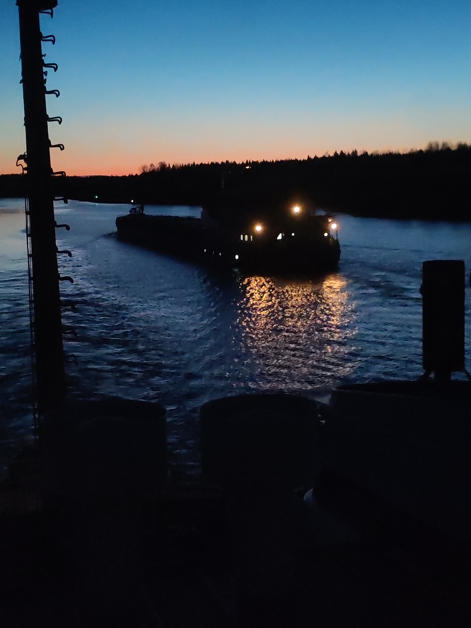 At dusk in Badozhskoi Pogost, Russia, a barge travels along a canal. The sky transitions from blue to orange near the horizon. Dark silhouettes of trees line the canal banks. The barge is illuminated by multiple lights, creating reflections in the water. In the foreground, a dark metallic structure with ladder rungs is positioned to the left, and a post is positioned to the right. Below these is dark indistinct machinery. The water shows the turbulence caused by the barge's passage.