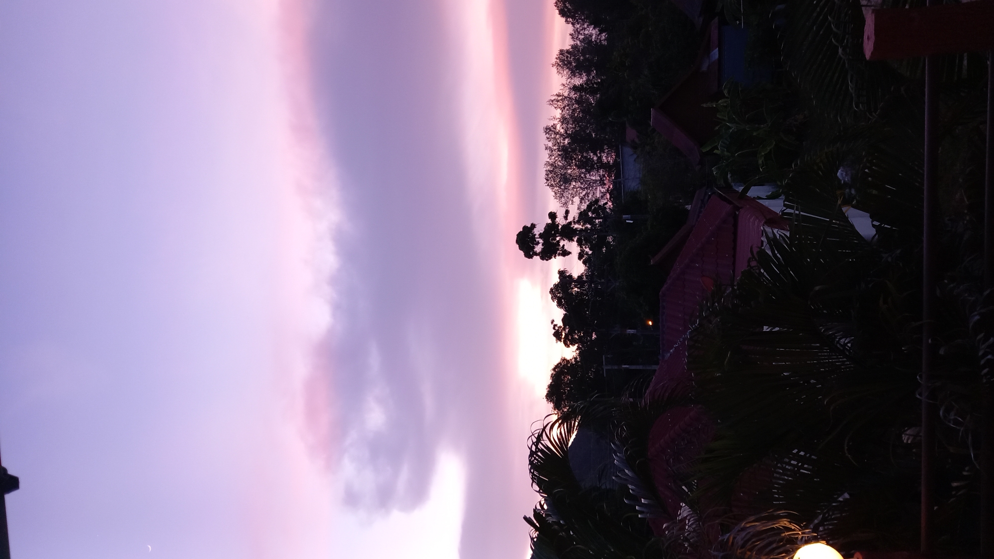 This is an image of a sunset or sunrise over a residential area in Ko Pha-ngan District, Thailand. The sky is a gradient of purple and pink, with a faint crescent moon visible in the upper left corner. Dark silhouettes of palm trees and other foliage are in the foreground, with red-tiled roofs of houses visible behind them. A single street light is also visible in the distance. The image is taken from a slightly elevated perspective, looking out over the neighborhood.