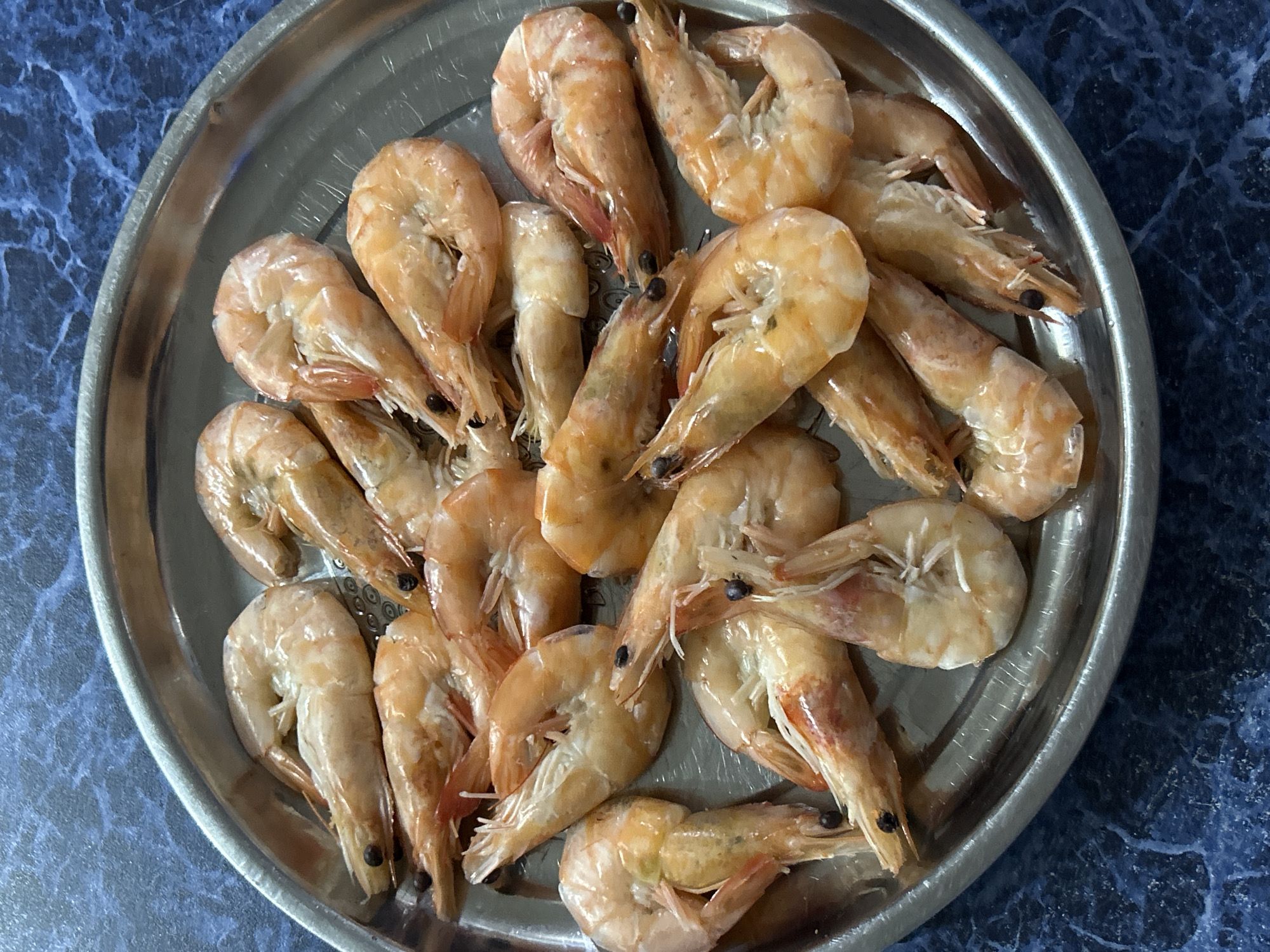 A metal plate is filled with cooked shrimp. The shrimp are pinkish-orange in color and appear to be arranged randomly on the plate. The plate sits on a blue marbled surface. The image does not contain any information relating to Rusalevka, Russia.