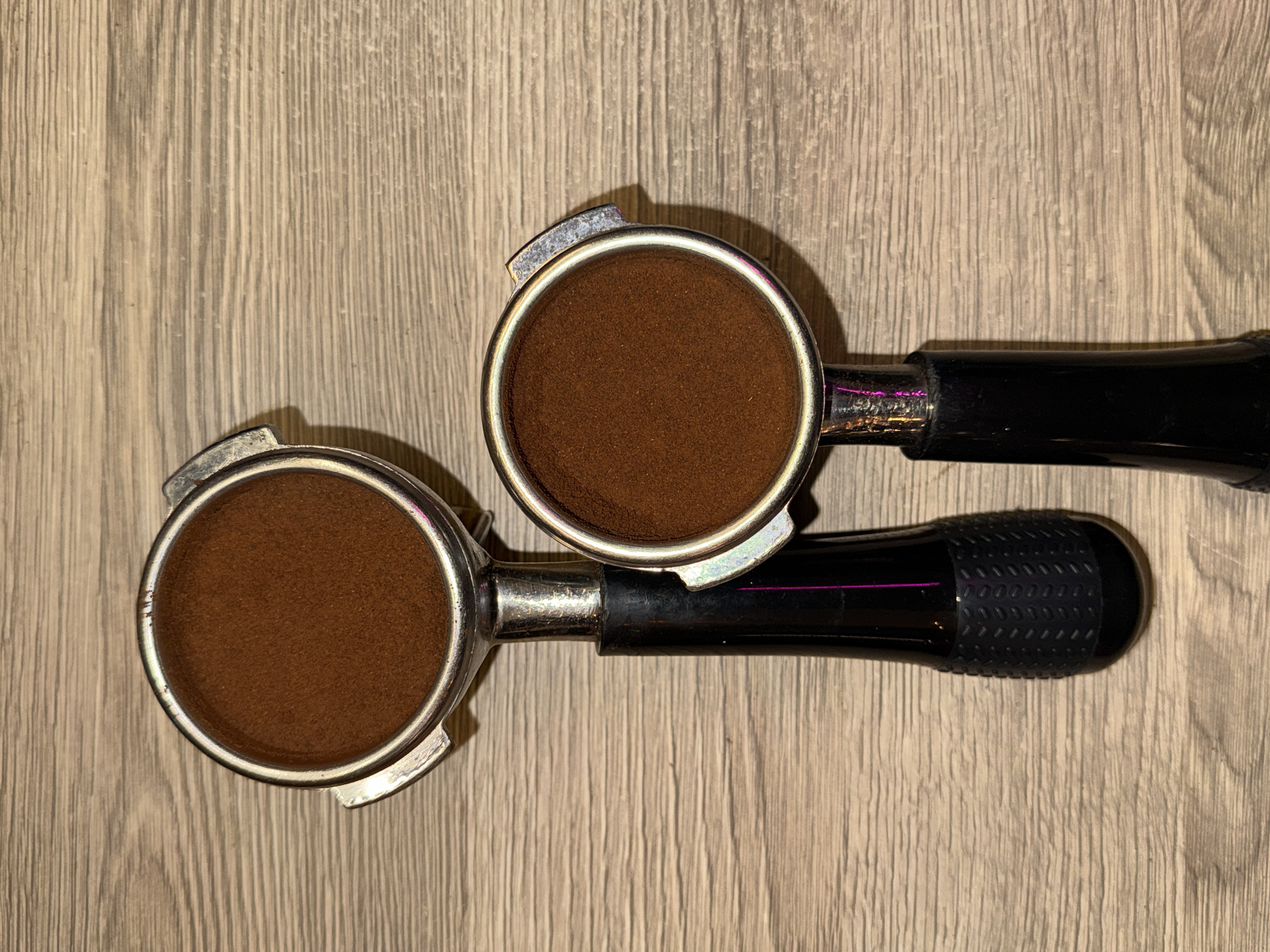The image shows two espresso machine portafilters filled with coffee grounds, lying on a wood-grain surface. The portafilters have silver metal heads and black plastic handles. One portafilter is slightly on top of the other, and the coffee grounds are visible within each filter basket. The surface appears to be a wooden tabletop or counter. There is no indication of the location being Dolgoprudny, Russia, or any people, actions, or interactions in the image.