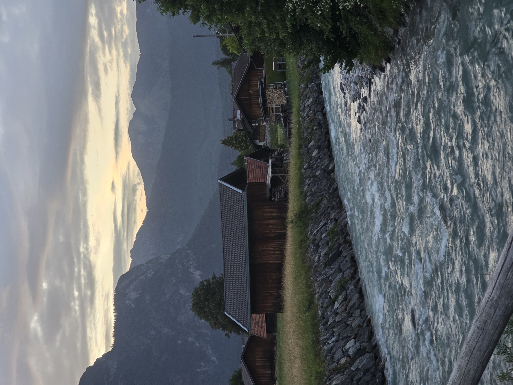 The image shows a pastoral scene with wooden buildings nestled at the foot of mountains, alongside a flowing river. The sky is overcast with hints of sunset colors. A river with visible currents and ripples flows from the foreground into the distance. The riverbanks are lined with rough stones and grass. Several traditional wooden barns and houses with dark-tiled roofs are visible on the grassy bank opposite the river. In the background, layered mountains rise, their peaks shrouded in mist or low clouds. The overall atmosphere is serene and natural, suggesting a rural or alpine setting. This scene is consistent with the landscapes found in and around Garmisch-Partenkirchen, Germany, a town known for its picturesque mountain scenery and traditional architecture. No people are visible in the image.