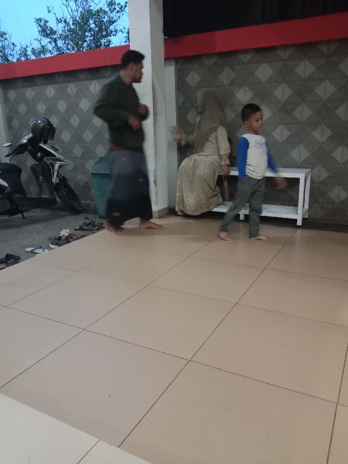 The image captures an open-air covered area in the city of Cabean, Indonesia, during twilight conditions. The foreground consists of light-colored square floor tiles. Three individuals are present: an adult male, an adult female, and a young male child. The adult male, standing on the left, is barefoot, dressed in a dark green long-sleeved shirt and a dark sarong, and is shown in profile, facing right, with his left hand slightly raised. Behind him, two motorcycles are parked; one is white and black with a black helmet on the seat, and a dark green plastic trash can is visible. The adult female is seated on a white, two-tiered bench in the mid-right, barefoot, wearing a light-colored patterned long dress and a beige headscarf (hijab); her left hand is blurred, indicating motion. The young male child is walking left across the frame on the right side, barefoot, wearing a white long-sleeved shirt with blue sleeves and grey trousers. The background features a grey wall with a diagonal geometric pattern, topped by a prominent red horizontal band. Several pairs of sandals and shoes are scattered on the ground to the left of the tiled area.