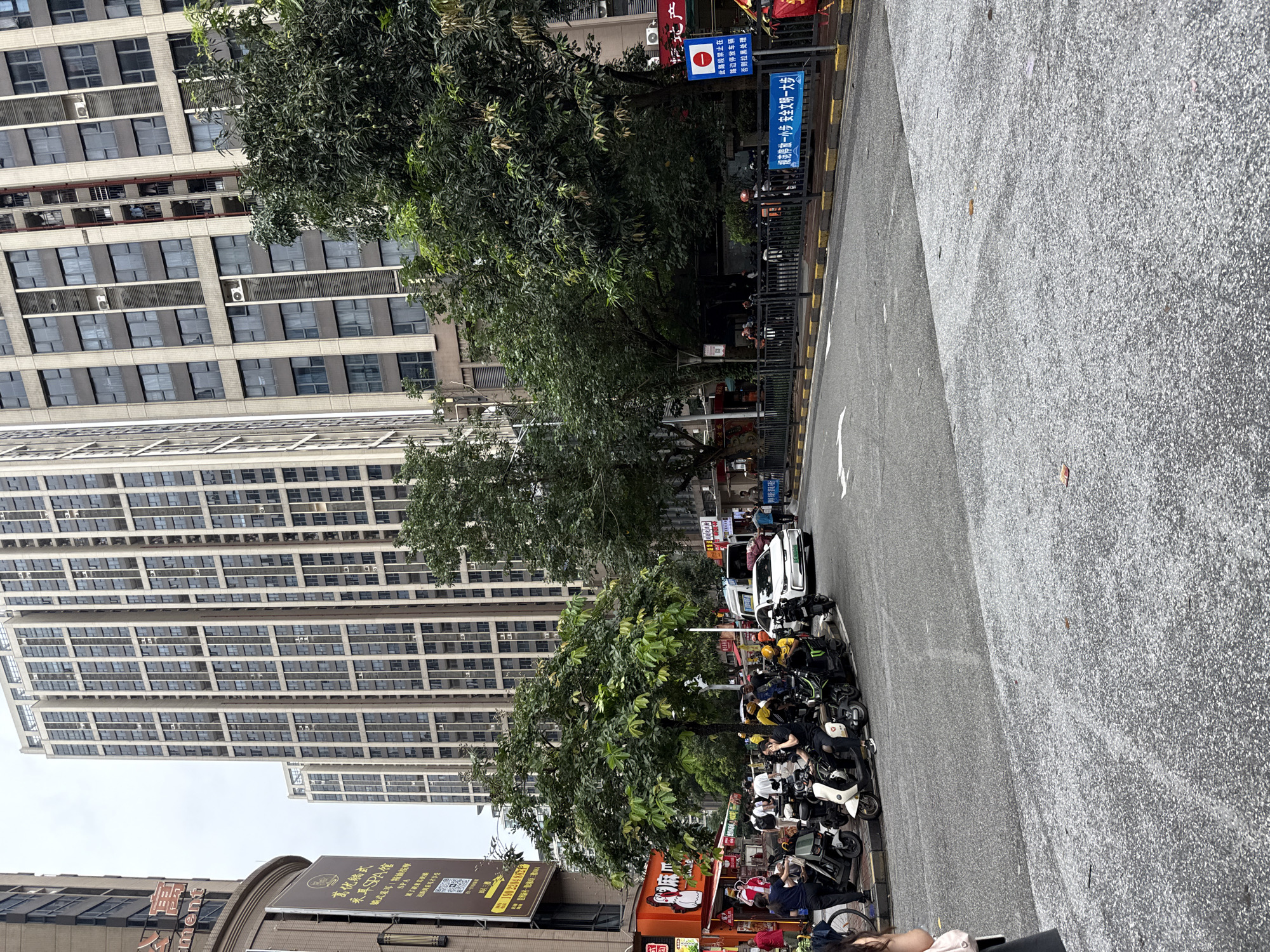 The image depicts a street scene in Haizhu District, China, characterized by modern high-rise buildings. In the foreground, a paved road is visible, with a line of parked electric scooters and several people interacting with them. A white car is also present on the road. Adjacent to the road, a black metal fence separates the sidewalk from a green, leafy area with trees, behind which more buildings are visible. Signage in Chinese characters is present on buildings and a blue signpost. The sky is overcast.