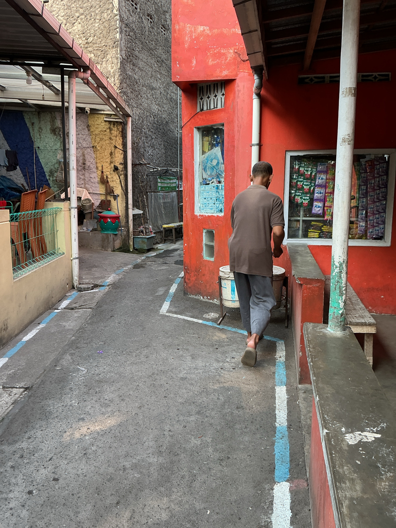 An outdoor, narrow, paved alleyway is depicted, likely in a residential area of Magelang, Indonesia. The alley is flanked by buildings. On the left, a light-colored wall with a green metal fence runs parallel to the alley. Beyond the fence, chairs and some tarpaulin are visible under a covered structure. The wall itself is decorated with diagonal stripes of blue, green, yellow, and pink.
On the right side of the alley, a vibrant red building dominates the scene. This building features at least one window with a lattice design and another window displaying various packaged goods, suggesting it might be a small shop or stall. A white downspout runs vertically down the red wall. A simple wooden bench is mounted against the red building.
In the center of the alley, a person is walking away from the camera. They are wearing a brown t-shirt and grey pants, with bare feet. The person is walking past two large cylindrical containers. The ground of the alley is paved with concrete and marked with white and blue lines. The overall impression is one of a lived-in, slightly worn urban environment.