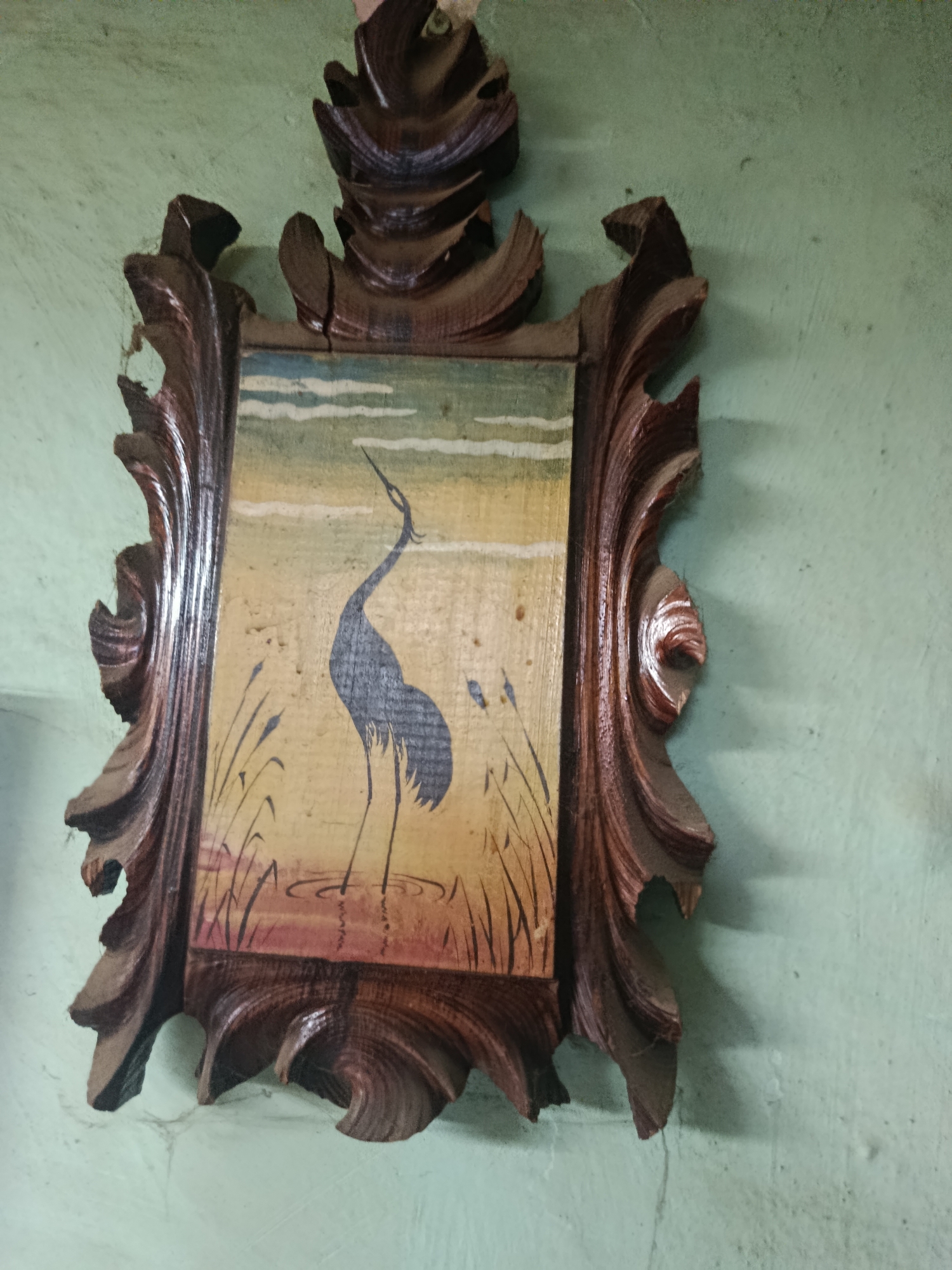 A framed painting of a heron in a marshy landscape hangs on a pale green, textured wall. The painting itself is of a dark blue heron standing in shallow water with reeds around it. The sky is a pale blue with white clouds. The frame is made of dark, carved wood and appears somewhat worn and damaged in places. There are no people visible in the image. The image does not contain any information about its location in Chișinău, Moldova.