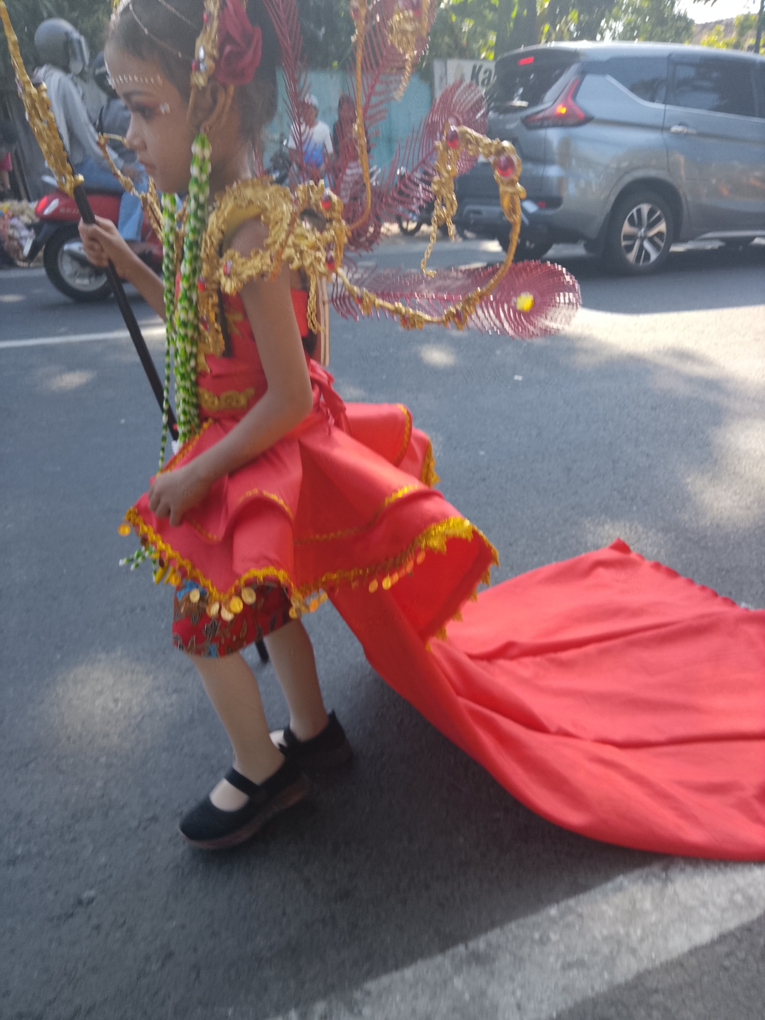 The image depicts a young girl in a traditional, elaborate red costume with gold embellishments, possibly participating in a parade or cultural event. She is holding a golden staff and has decorative wings attached to her back. Her costume features a short red dress with a flowing train, adorned with gold trim and coins. Underneath the dress, she wears patterned trousers. She is also wearing black Mary Jane shoes. The girl is captured in profile, looking to her left, with her face painted. The background shows a street scene with trees and vehicles, including a silver car and a red scooter, suggesting an outdoor urban environment. The lighting indicates it is daytime. The presence of what appears to be a parade suggests the location is Buduan, Indonesia, known for its vibrant cultural festivals.