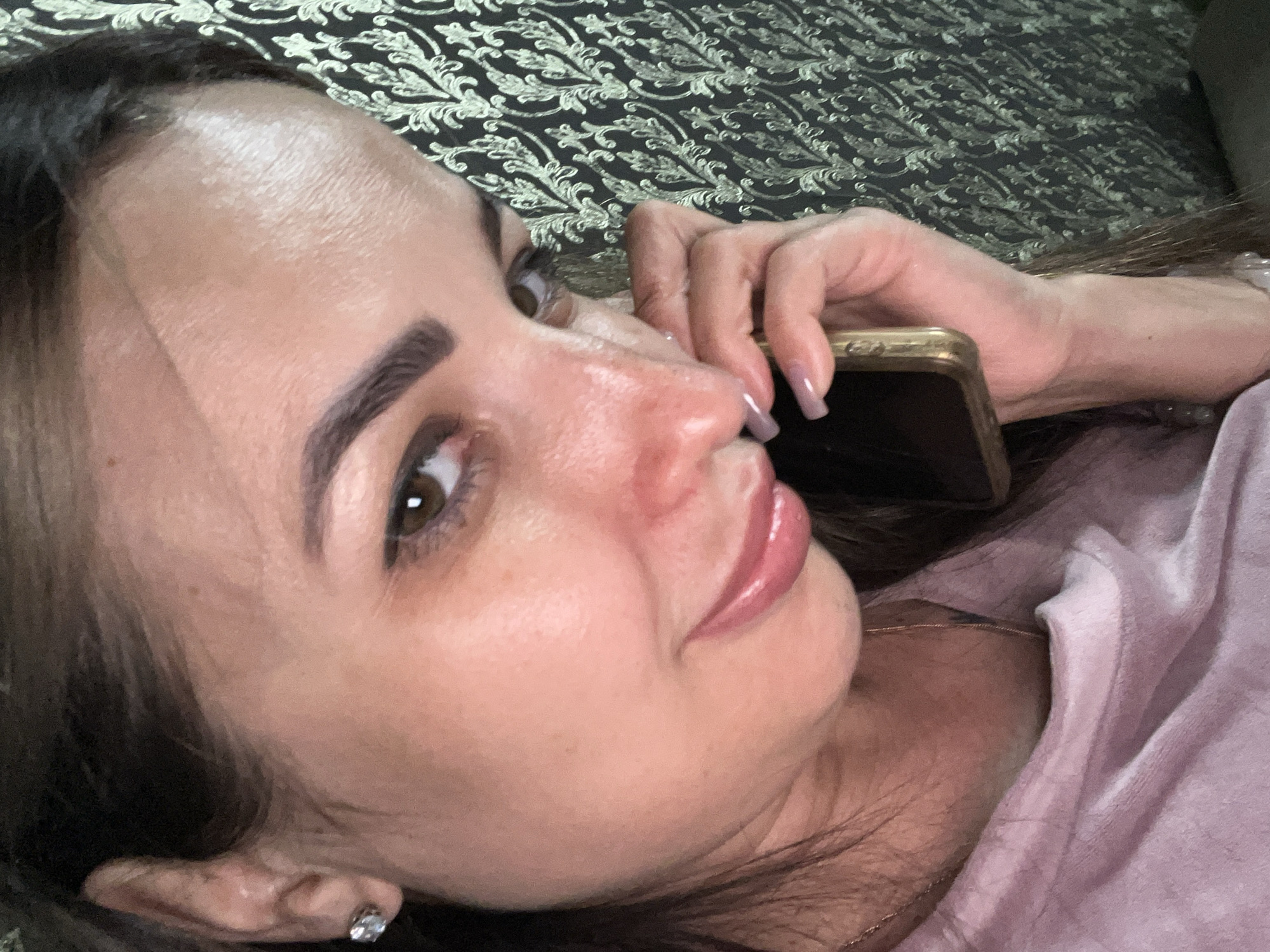 A close-up, over-the-shoulder selfie shows a woman holding a cell phone to her ear, looking away from the camera. The woman has dark hair, brown eyes, and is wearing a pink top and a diamond earring. The background is a dark, textured wallpaper with a floral pattern. The woman's face is partially illuminated by the phone screen.