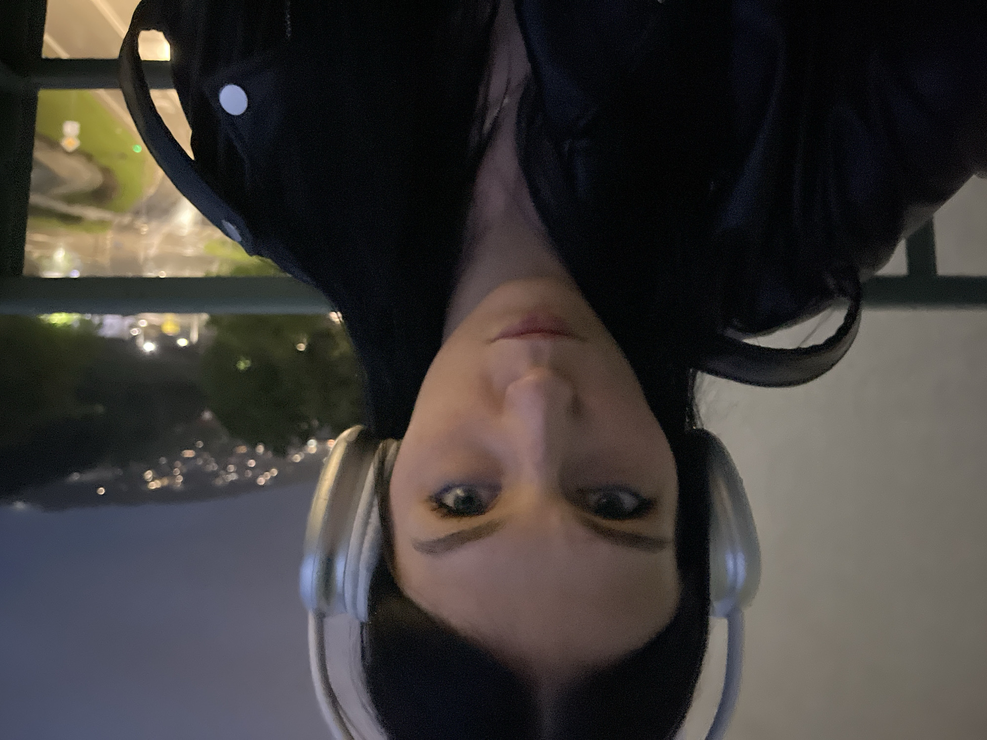 The image presents a close-up, upside-down portrait of a person, likely a young woman, captured at night. When correctly oriented, the woman's face is prominent in the upper half of the frame, looking directly into the camera with a neutral expression. She has light skin, dark hair, and noticeable light-colored eyes accented with eyeliner. She is wearing large, silver-toned over-ear headphones and a dark-colored jacket or top.
The background, visible behind and below her, is a blurred nightscape, viewed from an elevated position. A dark railing or barrier runs horizontally across the middle distance. Beyond this, numerous bokeh lights indicate an urban or suburban environment, possibly roads with vehicle lights or distant buildings. A few distinct green lights are visible, potentially traffic signals or other illuminated elements. The overall setting suggests a cool, dark evening outdoors, likely overlooking a city or town. No text is visible in the image.
