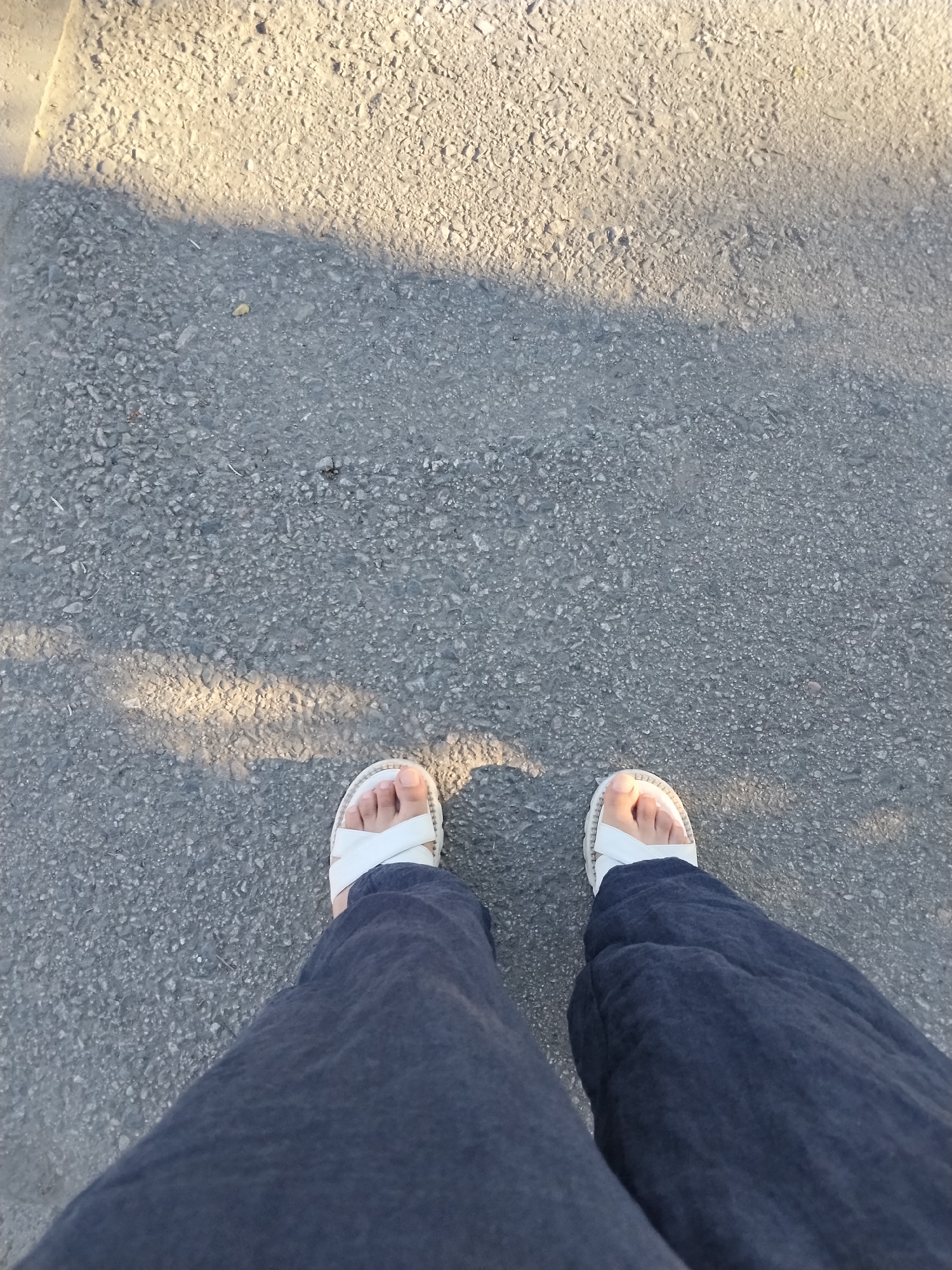 A first-person perspective shot looks down at a person's legs and feet as they stand on a gray asphalt surface. The person is wearing dark blue pants and white, cross-strap sandals. The asphalt is textured and appears to be a road or sidewalk. A diagonal shadow, possibly from a tree or building, falls across the asphalt, dividing it into a lighter and darker area. The image is taken outdoors, under natural lighting. The location is the city of Bishkek, Kyrgyzstan.
