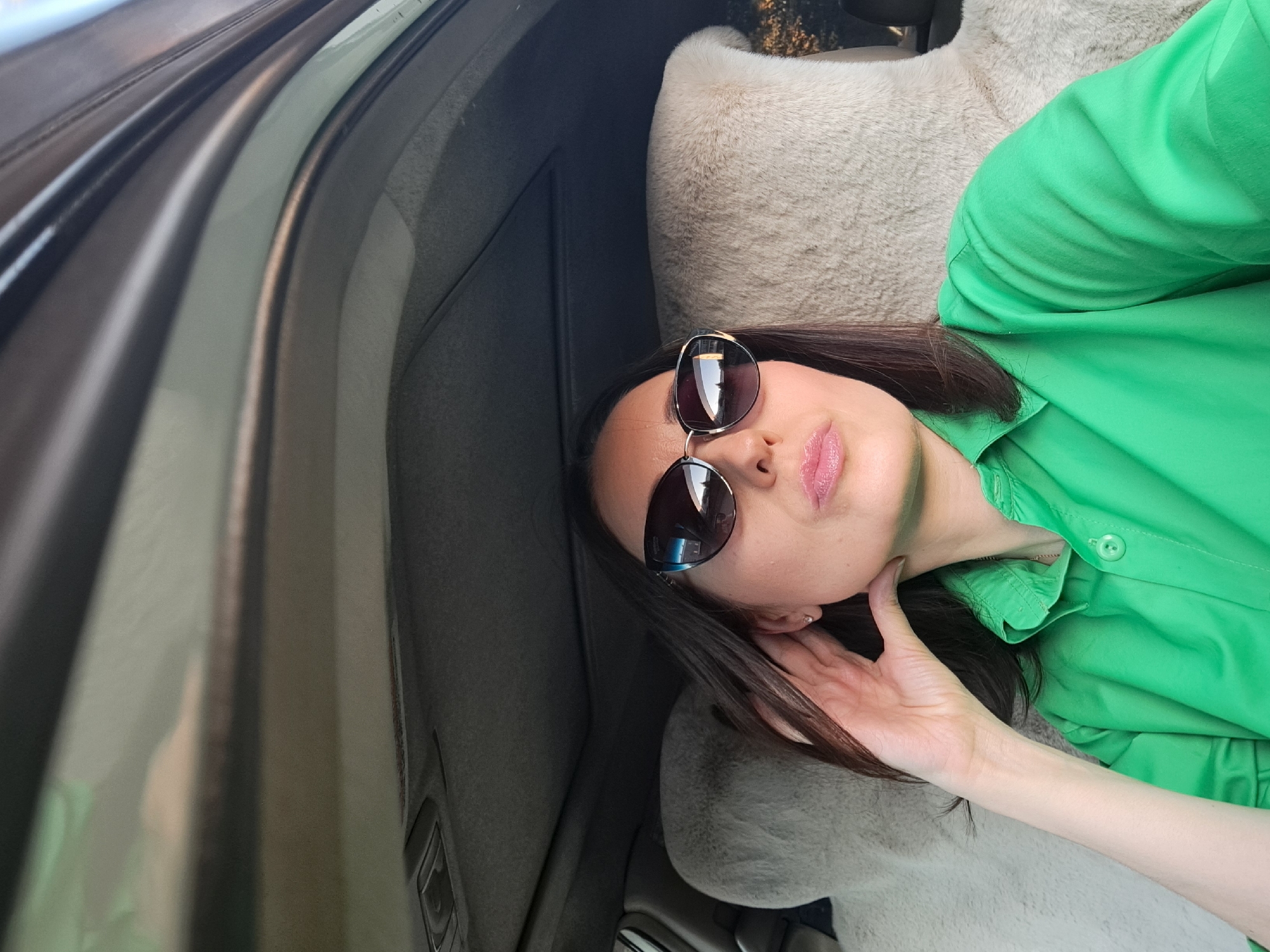 A woman with dark hair and sunglasses is sitting in the passenger seat of a car. She is wearing a green collared shirt. Her left hand is raised to her ear. The interior of the car is visible, including the headrests of the seats, which appear to be covered in a cream-colored fuzzy material, and the car's sunroof is open. The scene appears to be outdoors, possibly in Irkutsk, Russia, given the reflections in her sunglasses which suggest a cityscape with trees.