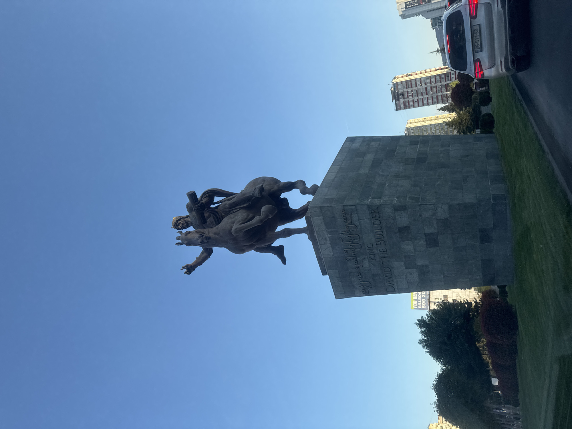 An equestrian statue of King David the Builder, depicted on horseback with an outstretched arm, stands prominently on a large, dark gray stone pedestal. The monument is set against a clear, bright blue sky, indicating sunny weather during daylight hours. Inscriptions on the pedestal are visible in both English and Georgian script, clearly stating "KING DAVID THE BUILDER."
The statue is situated in an urban environment, identified as Tbilisi, Georgia, with modern, multi-story buildings visible in the background. A strip of green grass separates the monument from a road or parking area. A white car, possibly an SUV, is partially visible from the rear, showing its license plate "BG-011-BB," which is a Serbian registration (Belgrade). Another notable detail includes a billboard on a distant building displaying partial text: "FOR REN BILLBOA 599 20." The scene captures a historical landmark within a contemporary cityscape.
