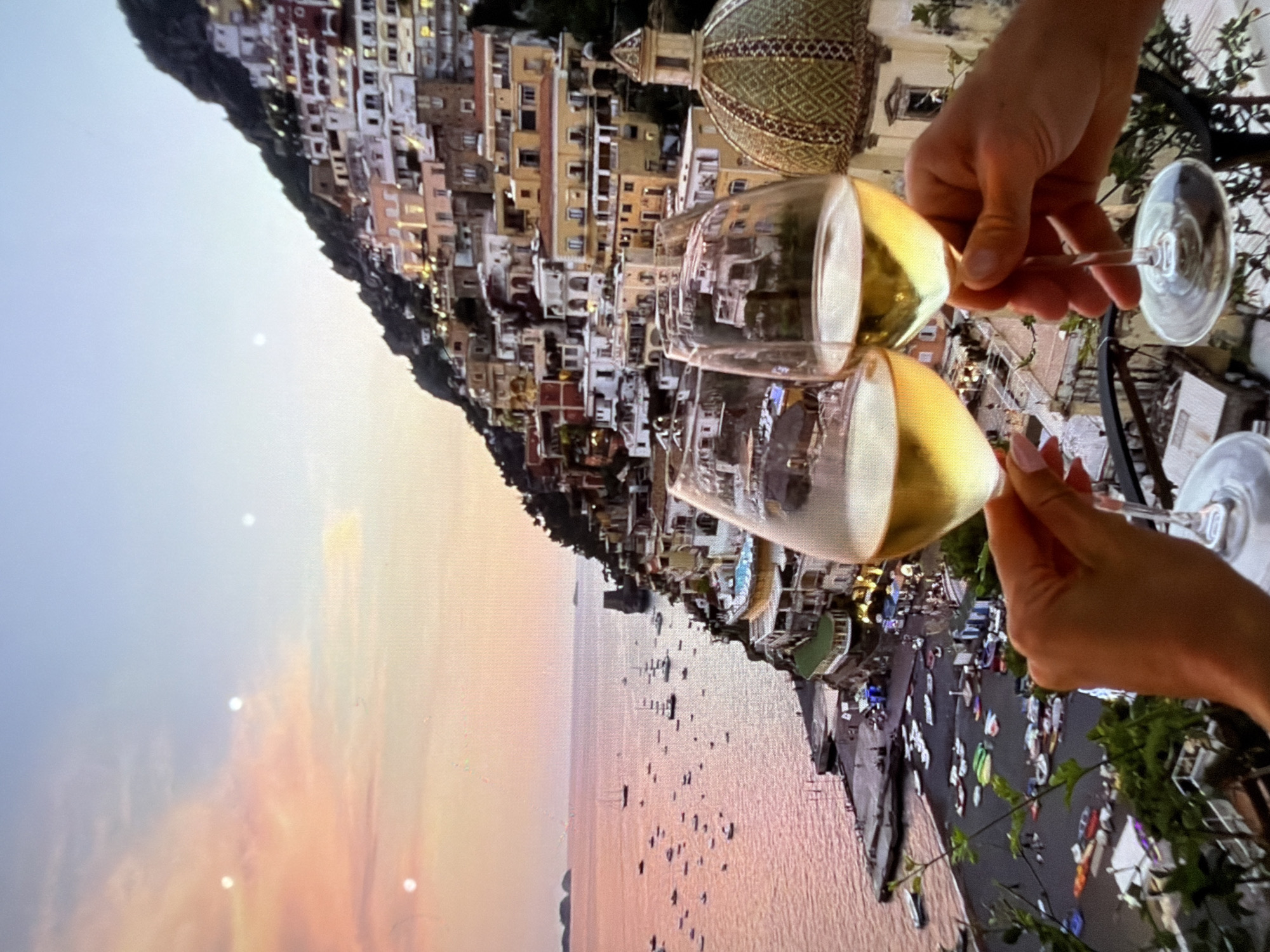 The image captures a scene from Positano, Italy. The view is framed by two hands holding wine glasses filled with a golden liquid. The background shows the city's distinct architecture, where buildings of various colors are stacked along the cliffside. Below, the sea stretches towards the horizon with numerous small boats dotting the water. The sky is a mix of hues, suggesting either sunrise or sunset. A dome-shaped structure with patterned tiles is visible among the buildings.