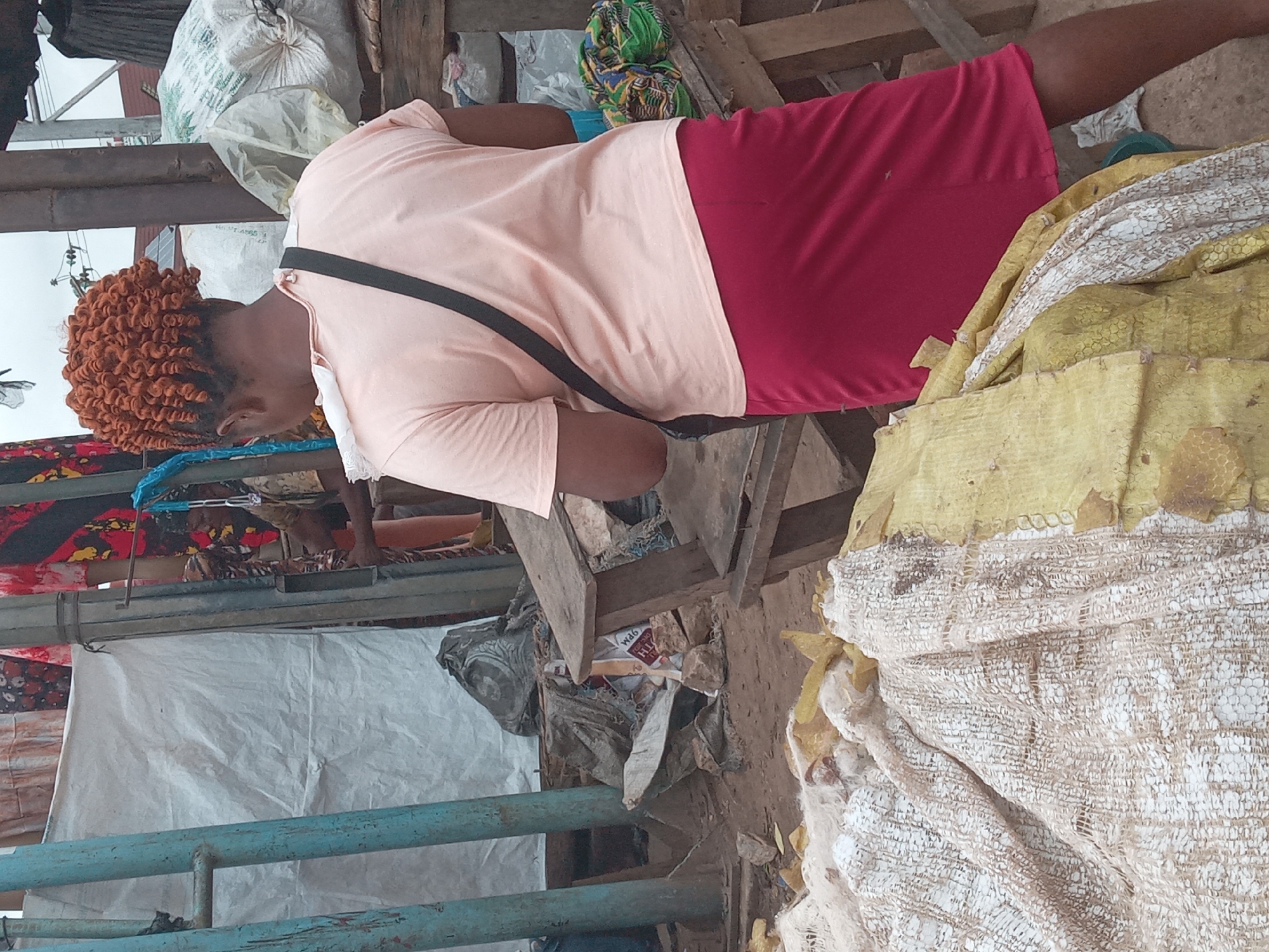 An individual with orange curly hair, a light pink t-shirt, and a maroon skirt stands among market debris in Onitsha, Nigeria. The person has a black strap across their shoulder and is looking down at something. The environment includes discarded textiles, metal scaffolding, bags of trash, and general market refuse. The scene is set in a crowded area, likely a marketplace due to the presence of textiles and other goods.