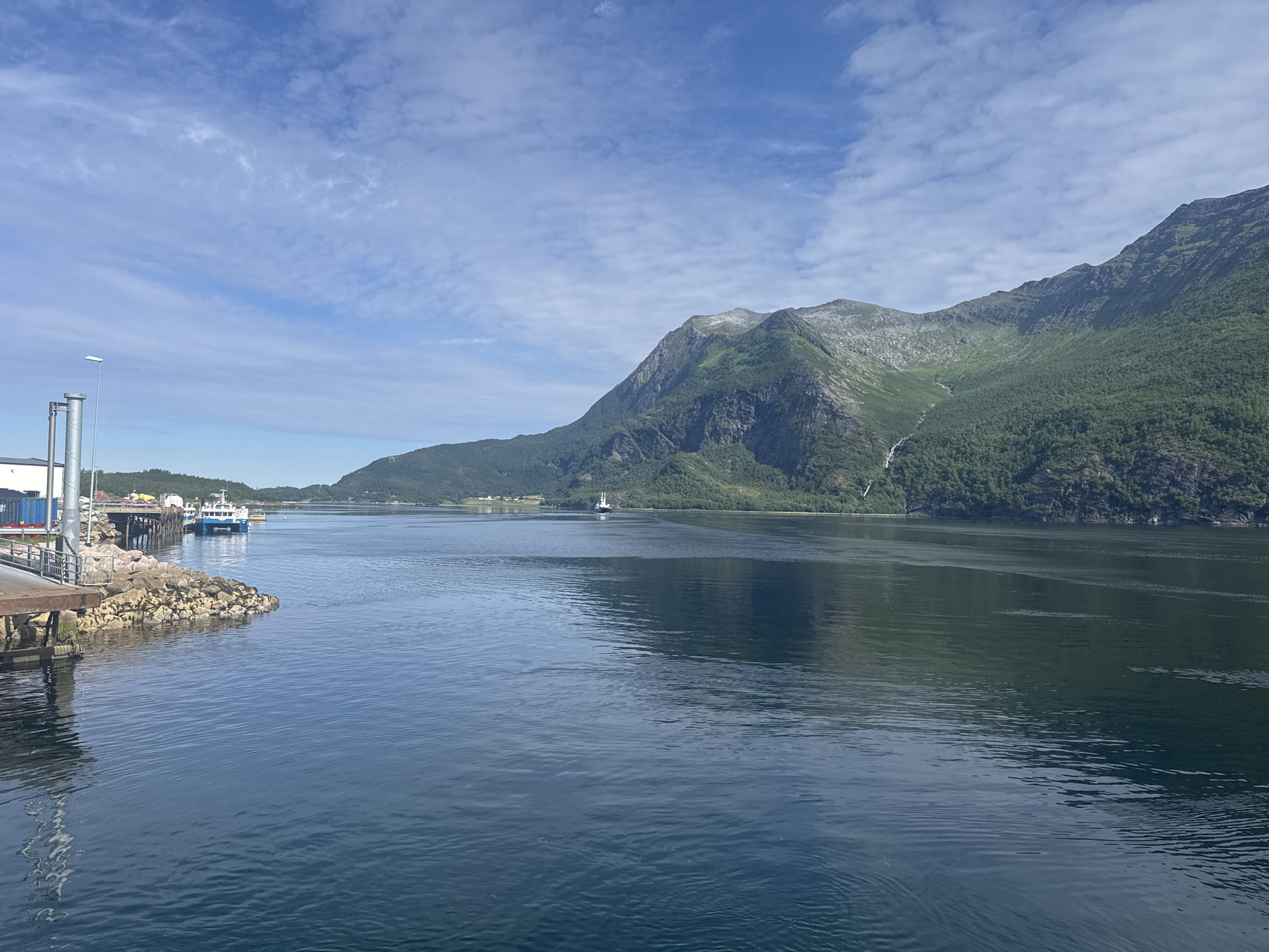 This is a photograph taken from a body of water looking towards a fjord in Inndyr, Norway. The scene depicts a calm, blue body of water with gentle ripples. In the distance, a large, green, mountainous landscape dominates the background, with some trees growing on its slopes. A narrow waterfall is visible cascading down the side of the mountain.
On the left side of the image, there is a pier with some structures, including containers and a lamppost. Two boats are docked at the pier. Further out in the water, a larger white ferry can be seen in the middle distance, moving away from the viewer. The sky is partly cloudy with a light blue hue. The overall impression is a serene and natural landscape, typical of a Norwegian fjord setting.
