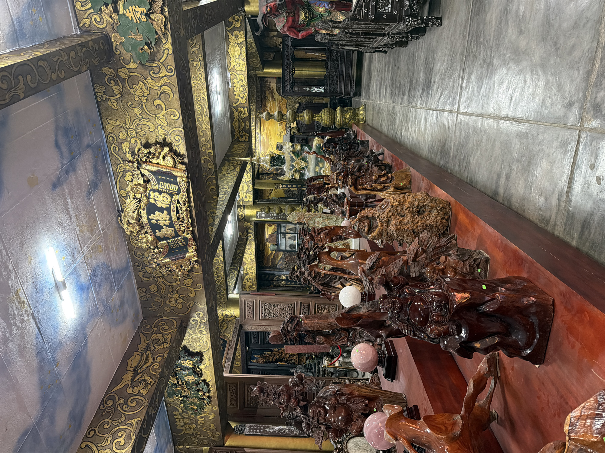 The image depicts the interior of a shop in Xuân Thành, Vietnam, filled with an extensive collection of carved wooden statues and decorative items. The items are displayed on red wooden shelves and tables, creating a rich, earthy color palette punctuated by the golden, ornate decorations on the walls and ceiling. The ceiling itself features a painted sky motif with clouds and stars, illuminated by fluorescent lights.
Prominently featured are numerous carved wooden figures, including depictions of deities, mythological creatures, and laughing Buddha statues. There are also large, smooth, pinkish spheres, possibly made of stone or crystal, placed on stands. In the background, more elaborate wooden furniture and intricate carvings are visible, along with a striking red-skinned, horned figure. The floor is a light gray, concrete-like material, marked by distinct seams. The overall impression is that of a well-stocked antique or craft shop specializing in traditional Asian art and decor.