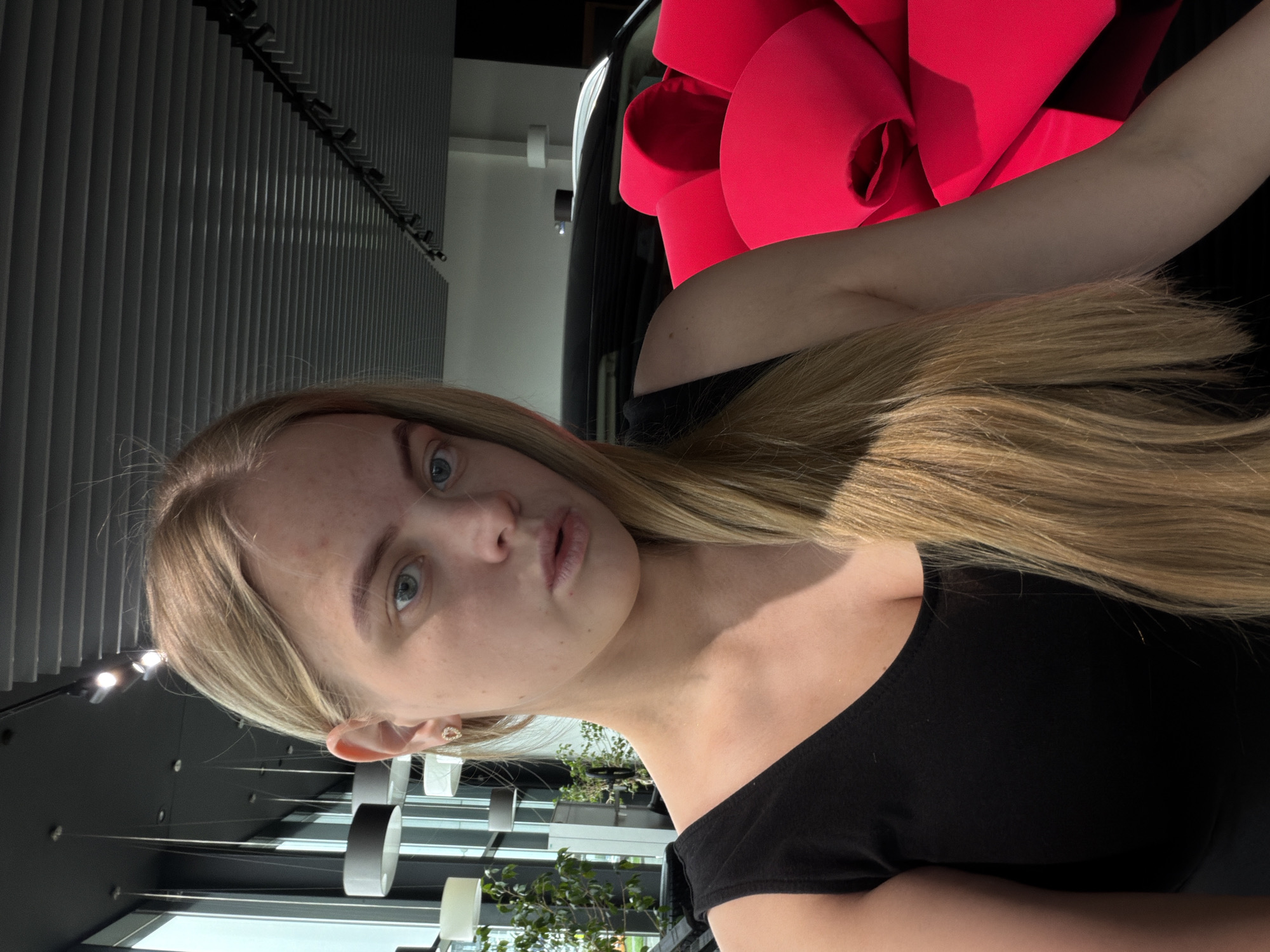 A fair-skinned young woman with blonde hair is depicted in a close-up shot. She is wearing a black off-the-shoulder top. Behind her is a car with a large red bow on it. The background includes a modern interior setting with visible ceiling fixtures and a window showing greenery. The location cannot be determined to be in Izhevsk, Russia from the image provided.