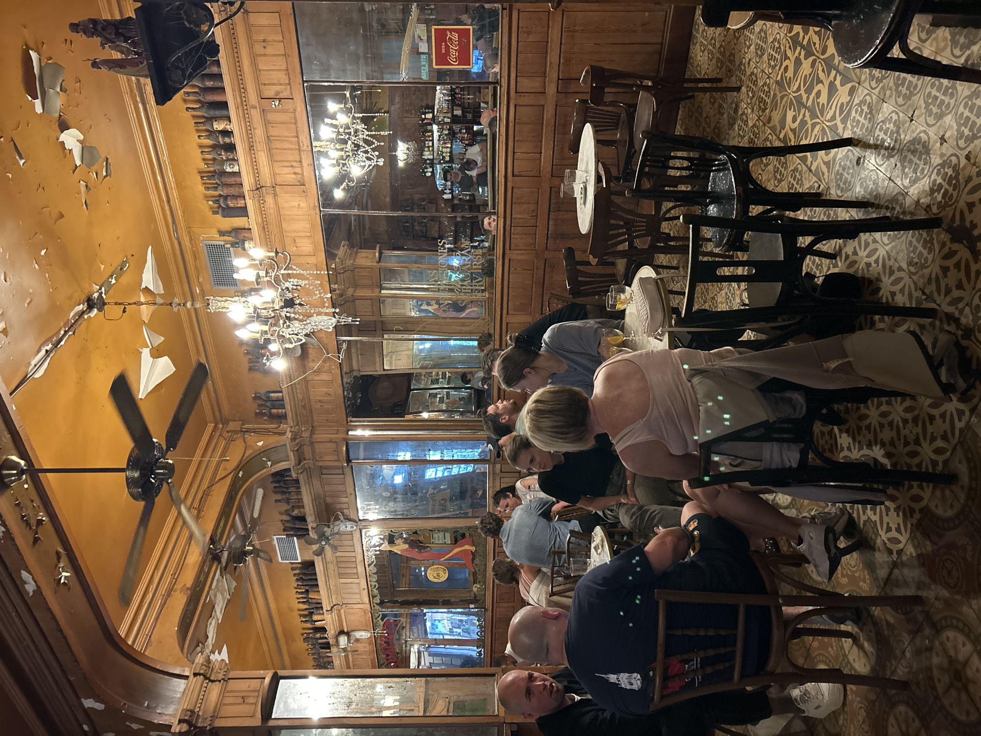 The image depicts a bustling interior scene, likely a restaurant or cafe, in Barcelona, Spain. The establishment is characterized by its traditional decor, featuring wooden panels, mirrored walls, and decorative lighting fixtures, including ornate chandeliers. Rows of bottles line the walls near the ceiling. The ceiling has peeling paint and several ceiling fans.
Several people are seated at tables, engaged in conversation and dining. The people are dressed in casual clothing. The flooring is patterned with geometric designs. The overall ambiance suggests a lively and historic atmosphere.
