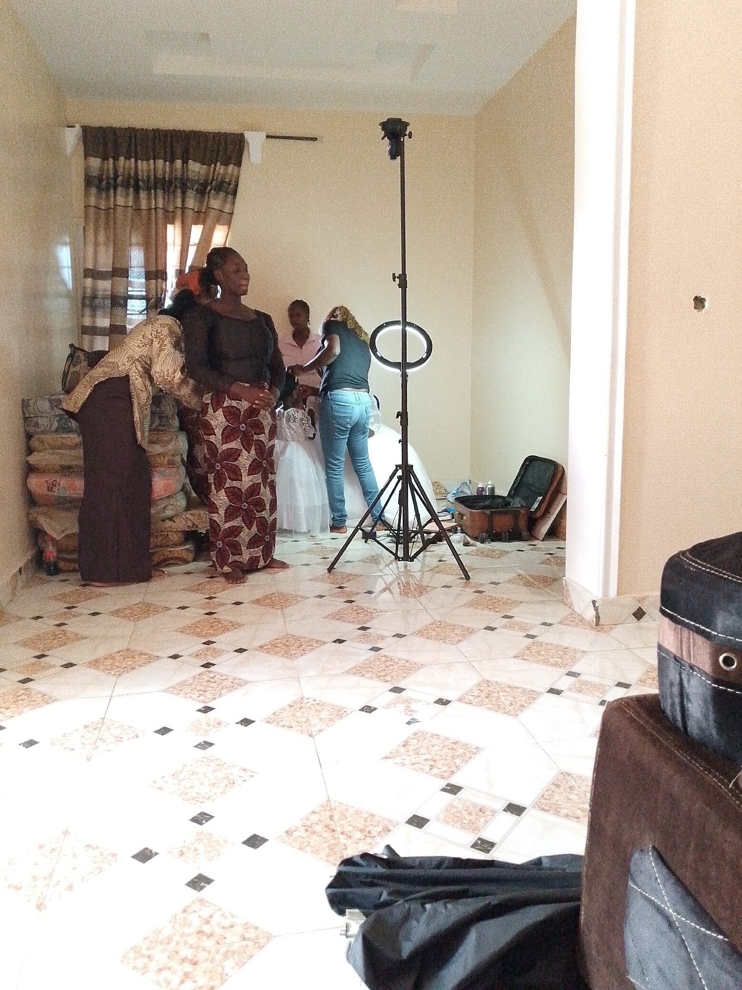 The interior of a room is depicted, possibly in an unknown location in the city of Unknown, Unknown. The room has tiled floors and cream-colored walls. To the left, there is a stack of cushions near a window covered with a brown patterned curtain. Four adults are visible. One woman is wearing a black top and a long patterned skirt. Another woman is wearing a gold-colored top and a long brown skirt. Another is wearing a pink shirt. The fourth woman is wearing a dark shirt and jeans. A young girl in a white dress is also present. There is a ring light on a stand and an open brown suitcase.